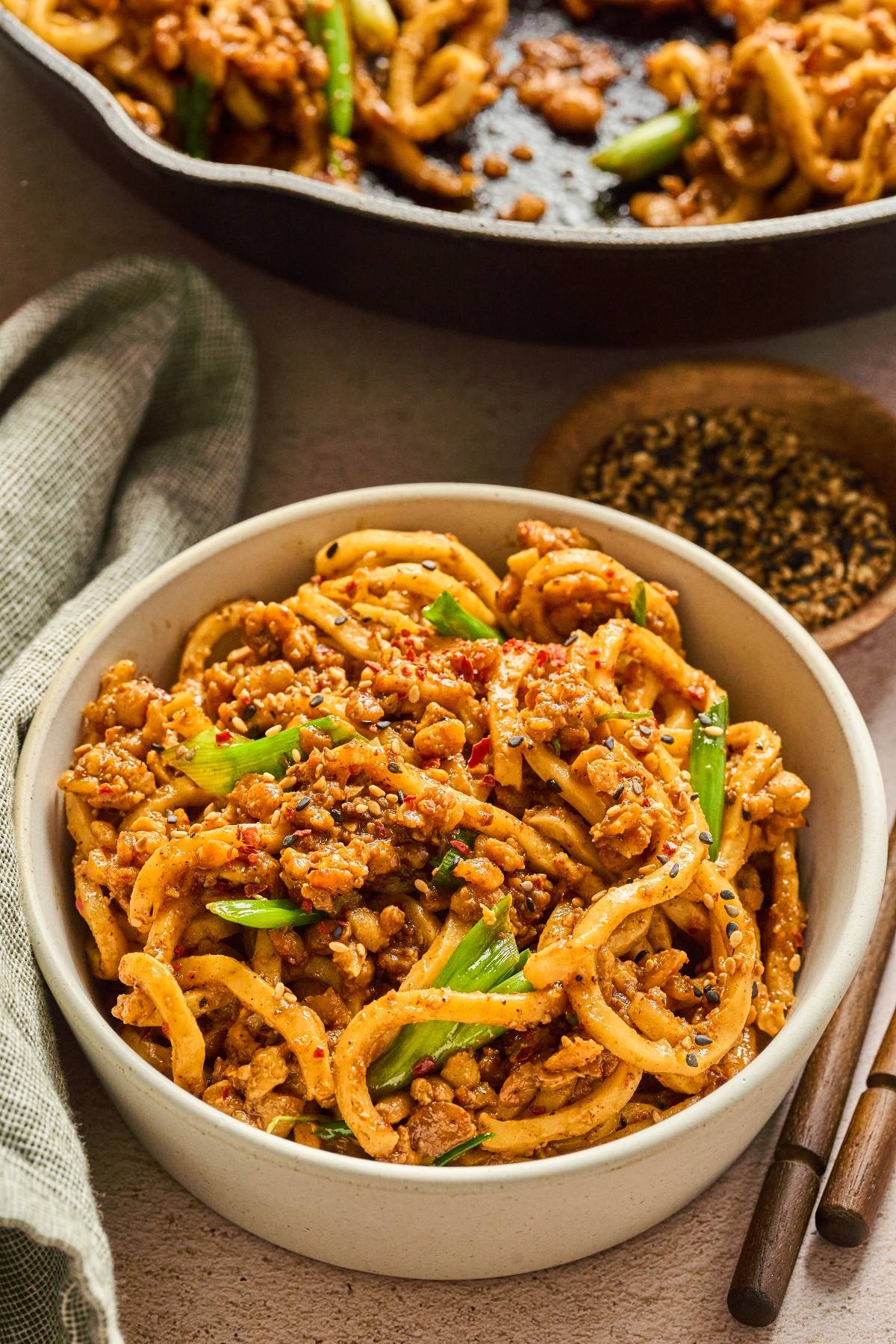 A bowl of spicy noodles with ground meat, green onions, and sesame seeds, served with chopsticks.