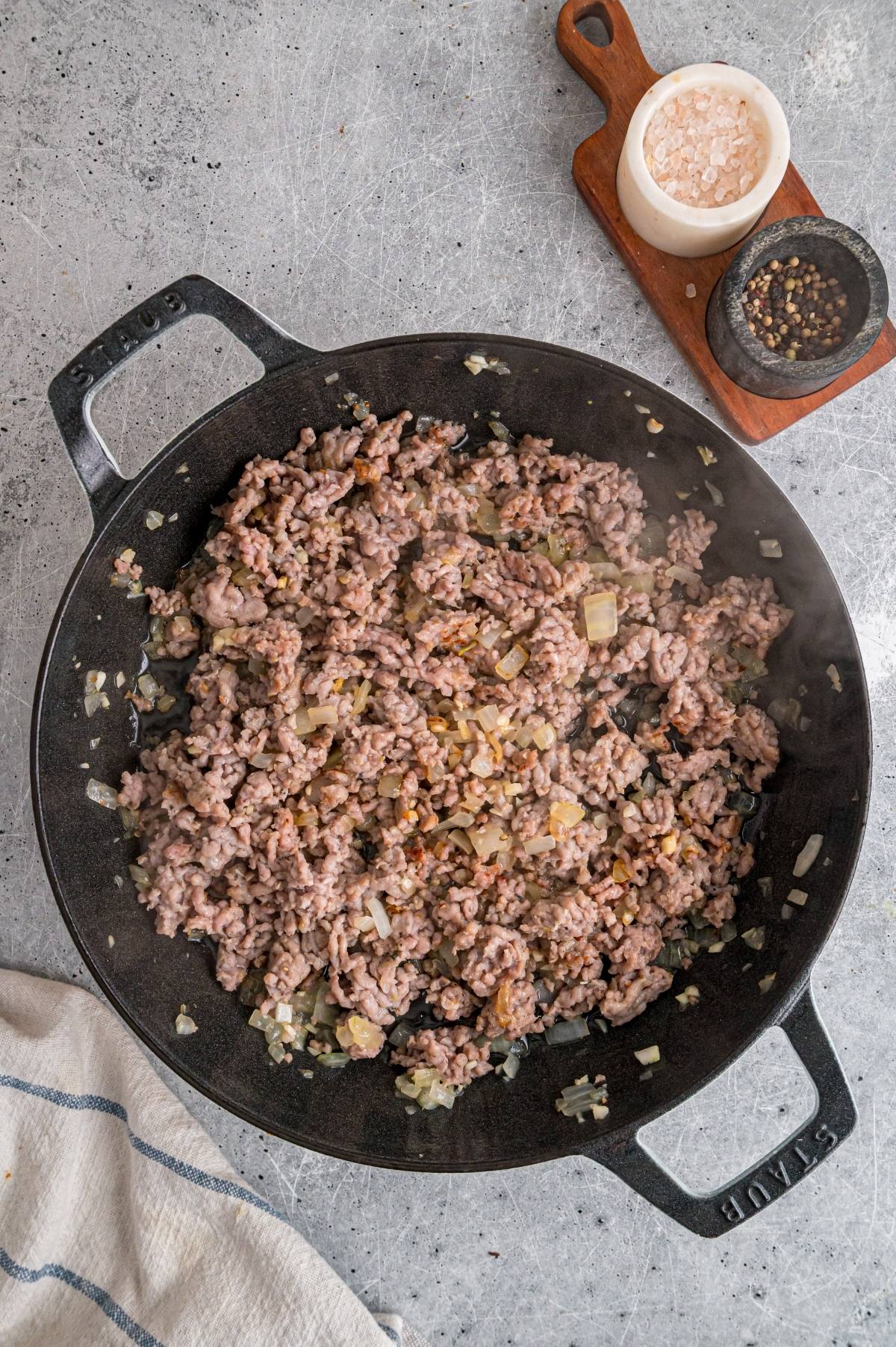 Ground beef and chopped onions cooking in a black skillet, with salt and pepper nearby on a wooden tray.