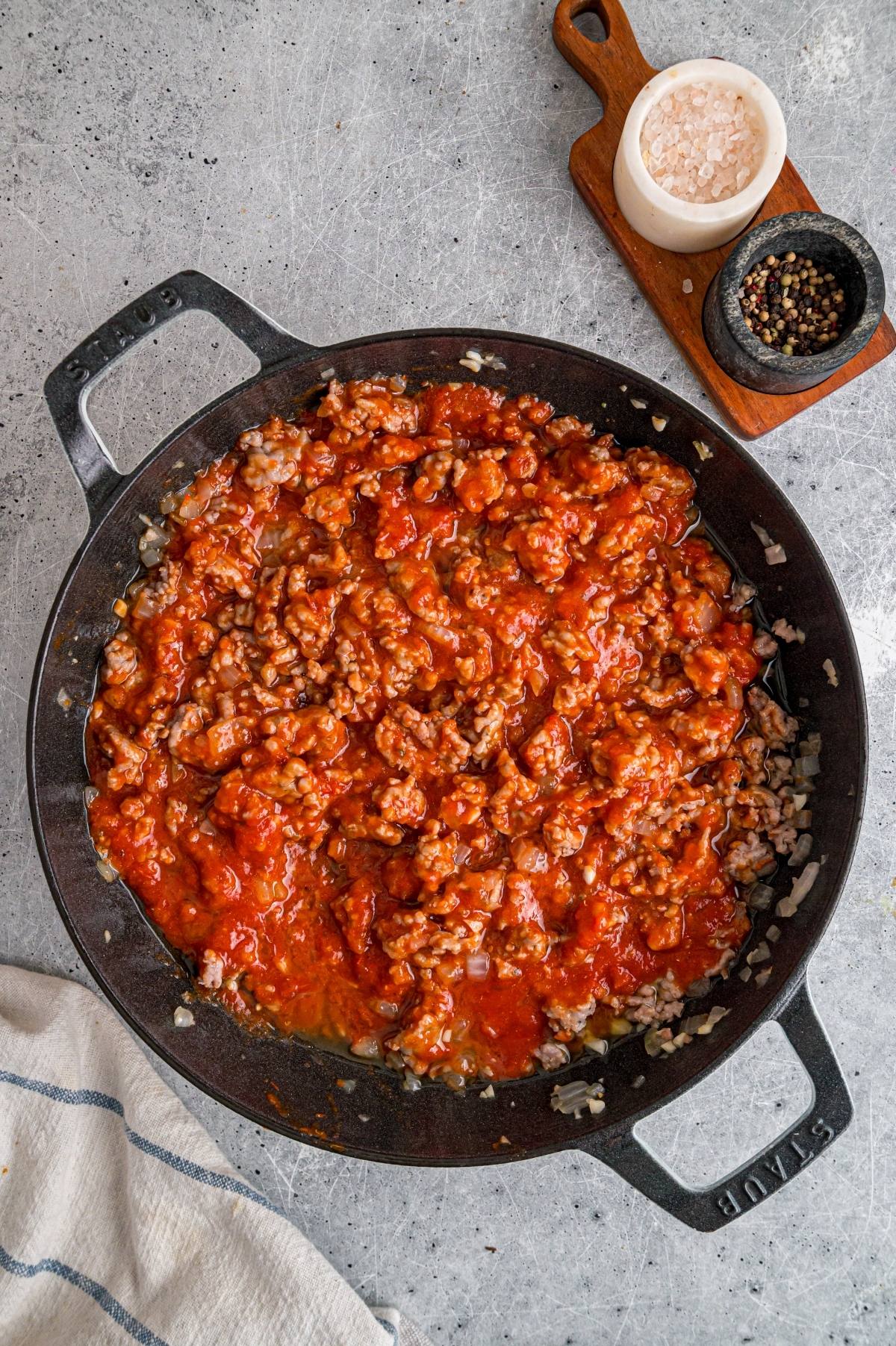 A pan filled with ground meat and tomato sauce sits on a gray surface near salt and pepper shakers.