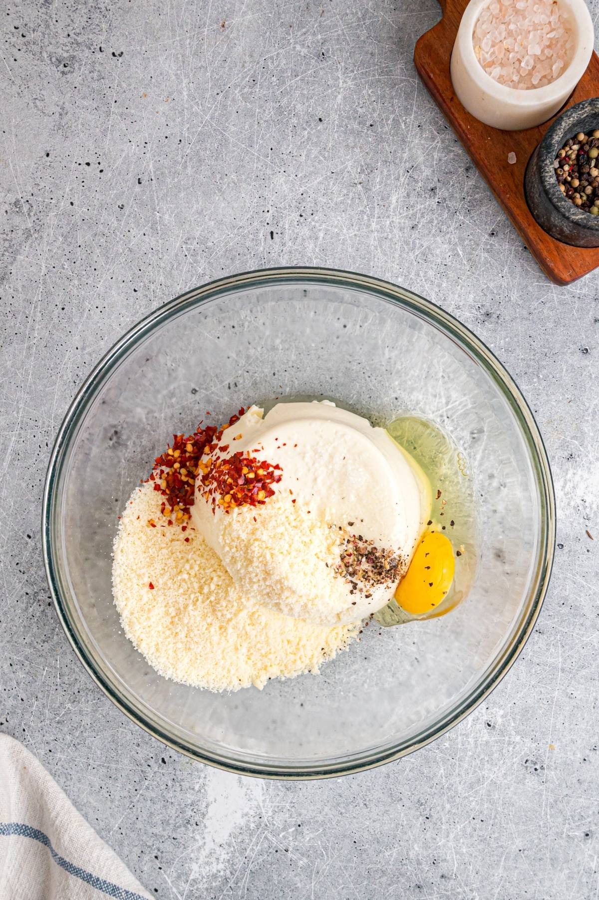 A glass bowl with ricotta, grated cheese, an egg, pepper, and chili flakes on a gray countertop.