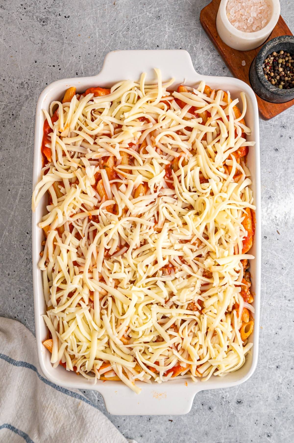Unbaked casserole dish filled with pasta, tomato sauce, and topped with shredded cheese on a gray surface.