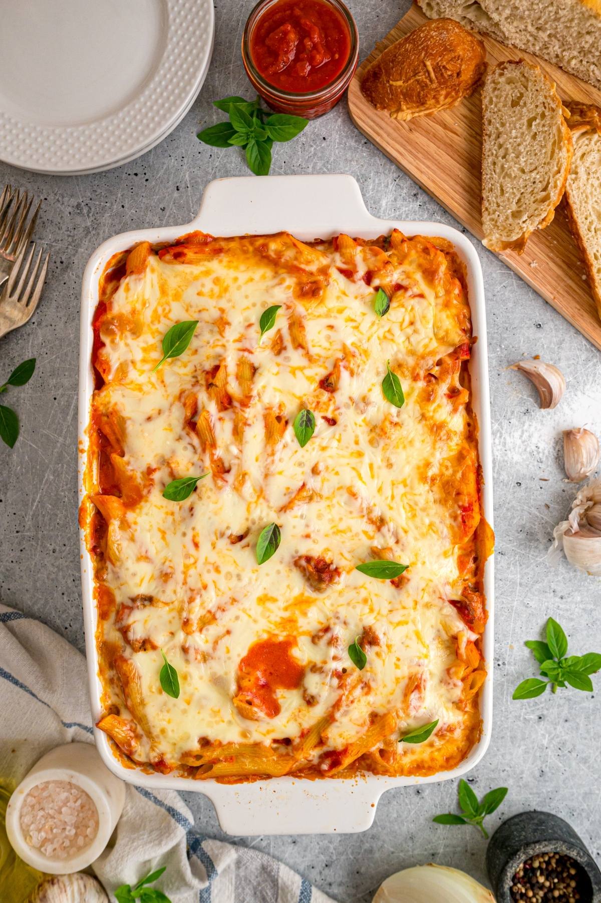 Baked mostaccioli pasta topped with melted cheese and basil in a white dish, surrounded by bread and sauce on a table.