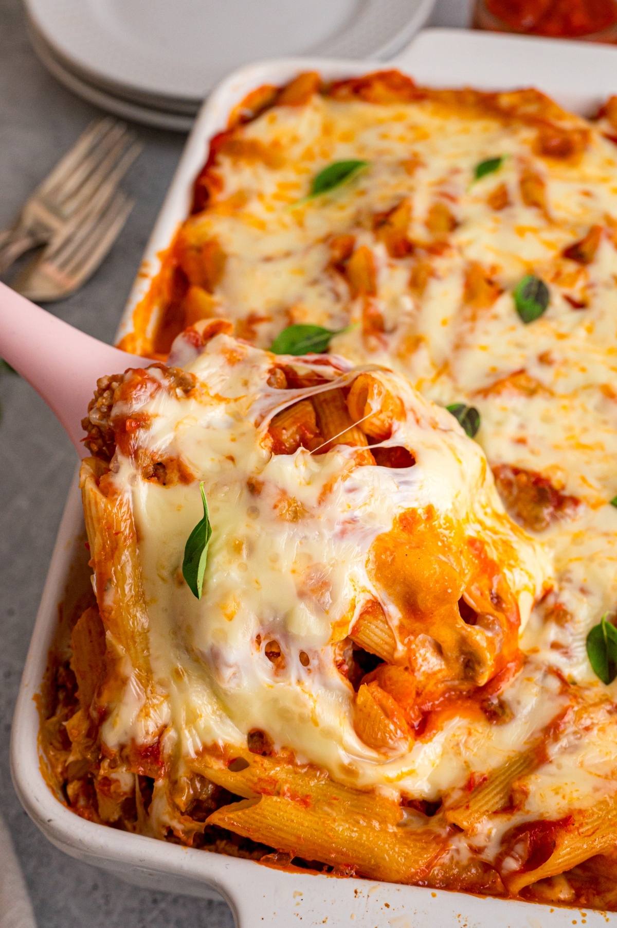 A cheesy baked pasta dish is being served from a casserole dish, topped with melted cheese and basil leaves.