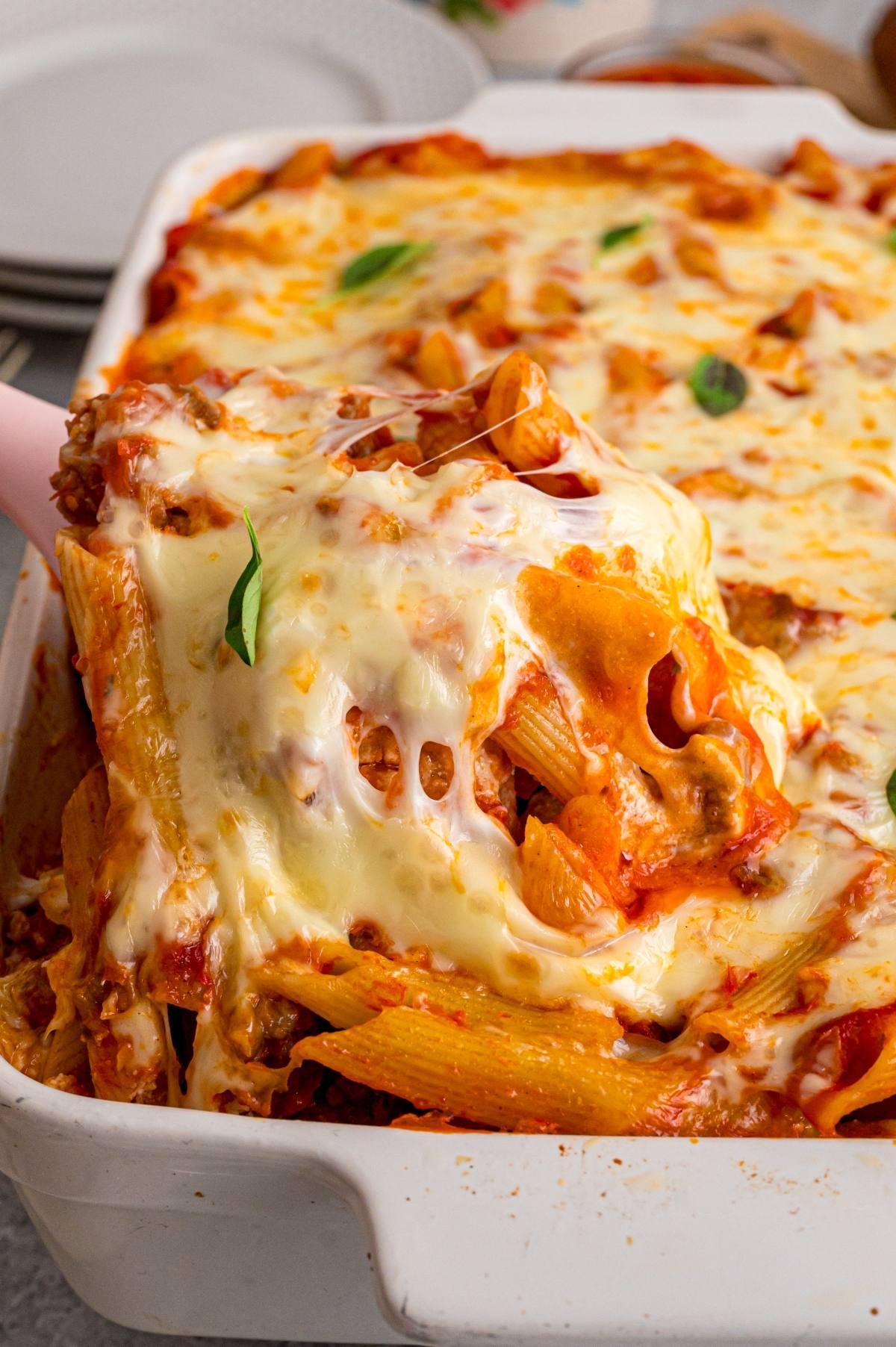 A cheesy baked pasta dish is being served from a white casserole dish with a serving spoon.
