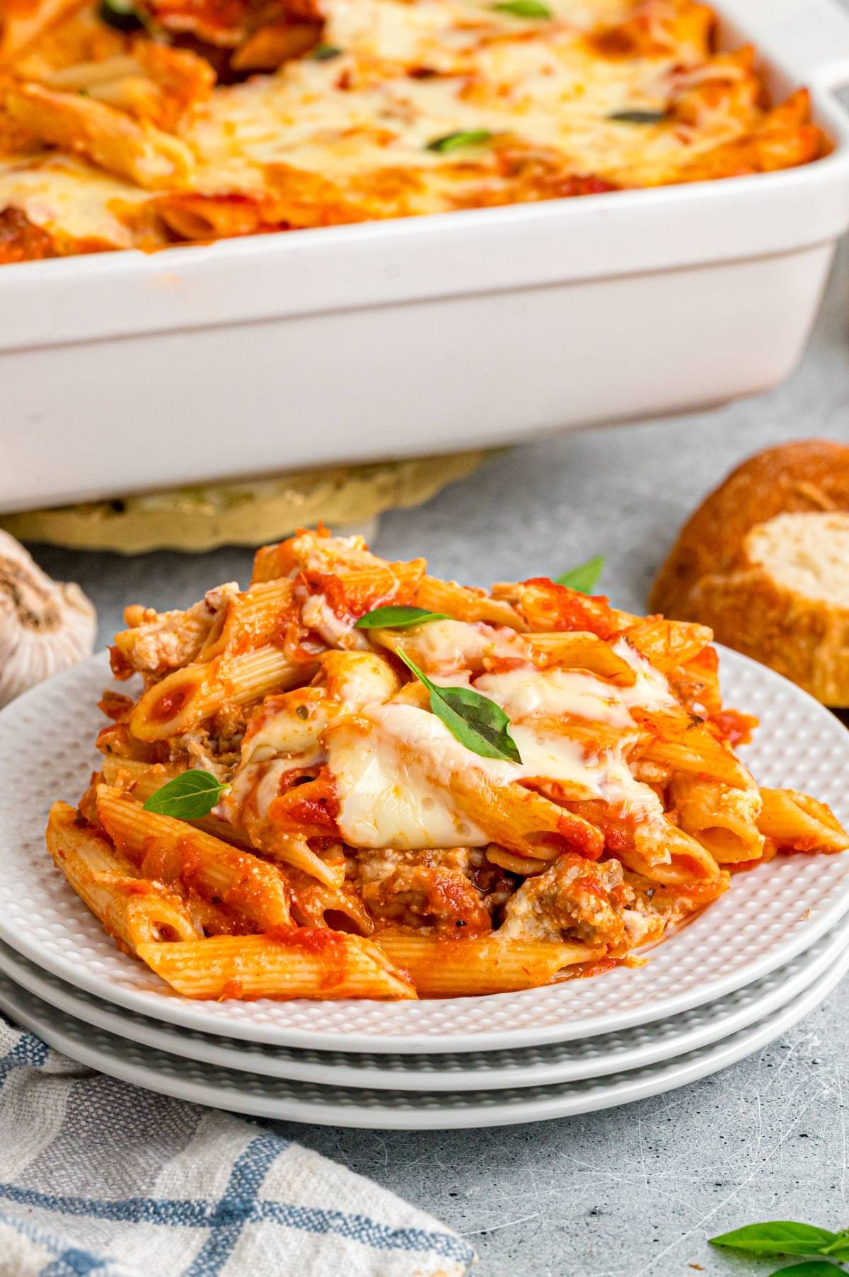A serving of cheesy baked mostaccioli pasta with tomato sauce on a plate, with a casserole dish and bread in the background.