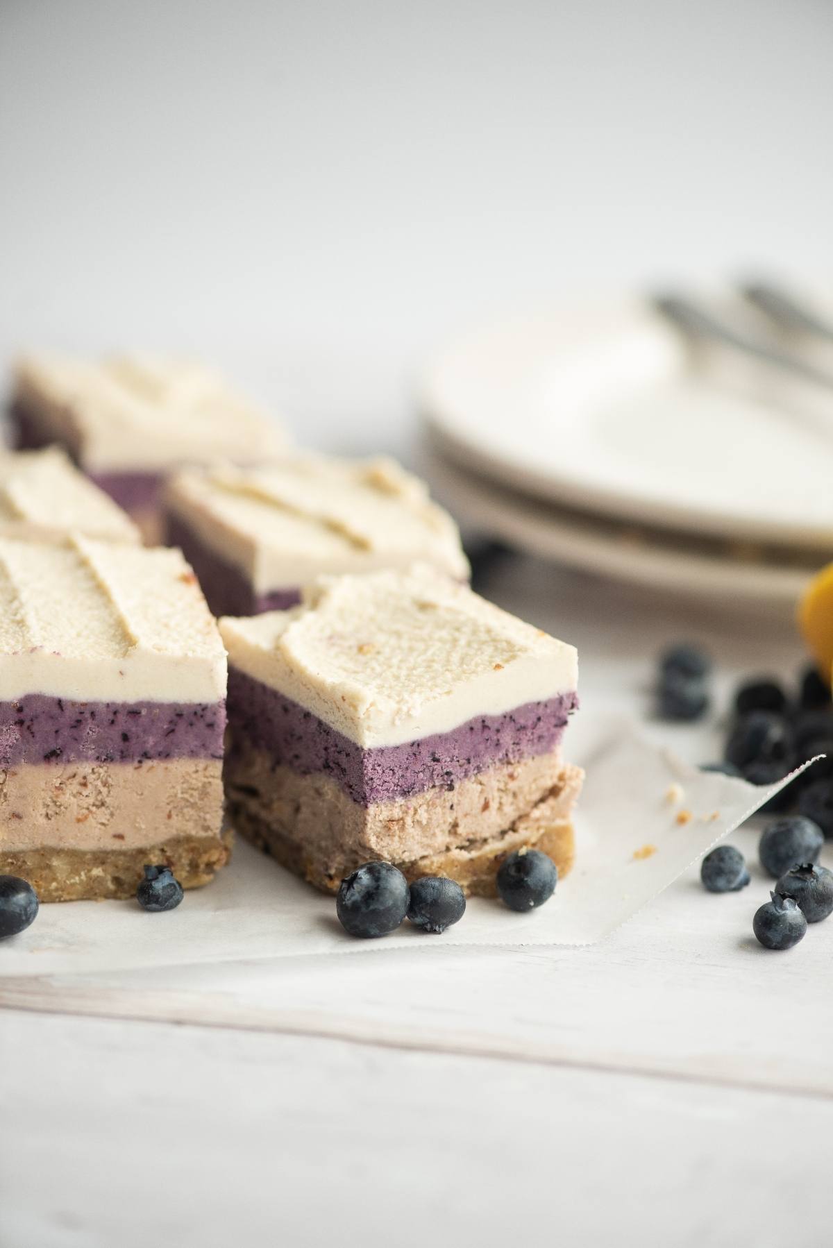 No Bake Berry Layered Bars featuring three layers and juicy blueberries, made with raw cashews for a creamy texture. Pictured on parchment paper with plates and forks in the background—a delicious no bake vegan cheesecake treat.
