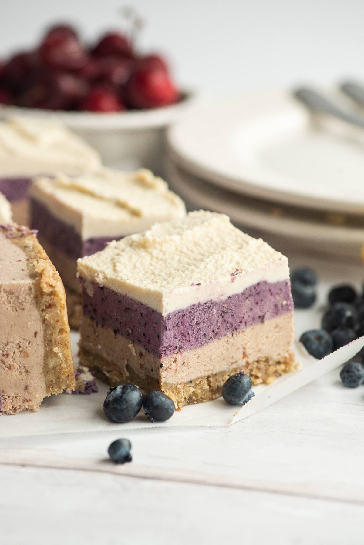 A square slice of no bake vegan cheesecake with layers and fresh blueberries scattered nearby.