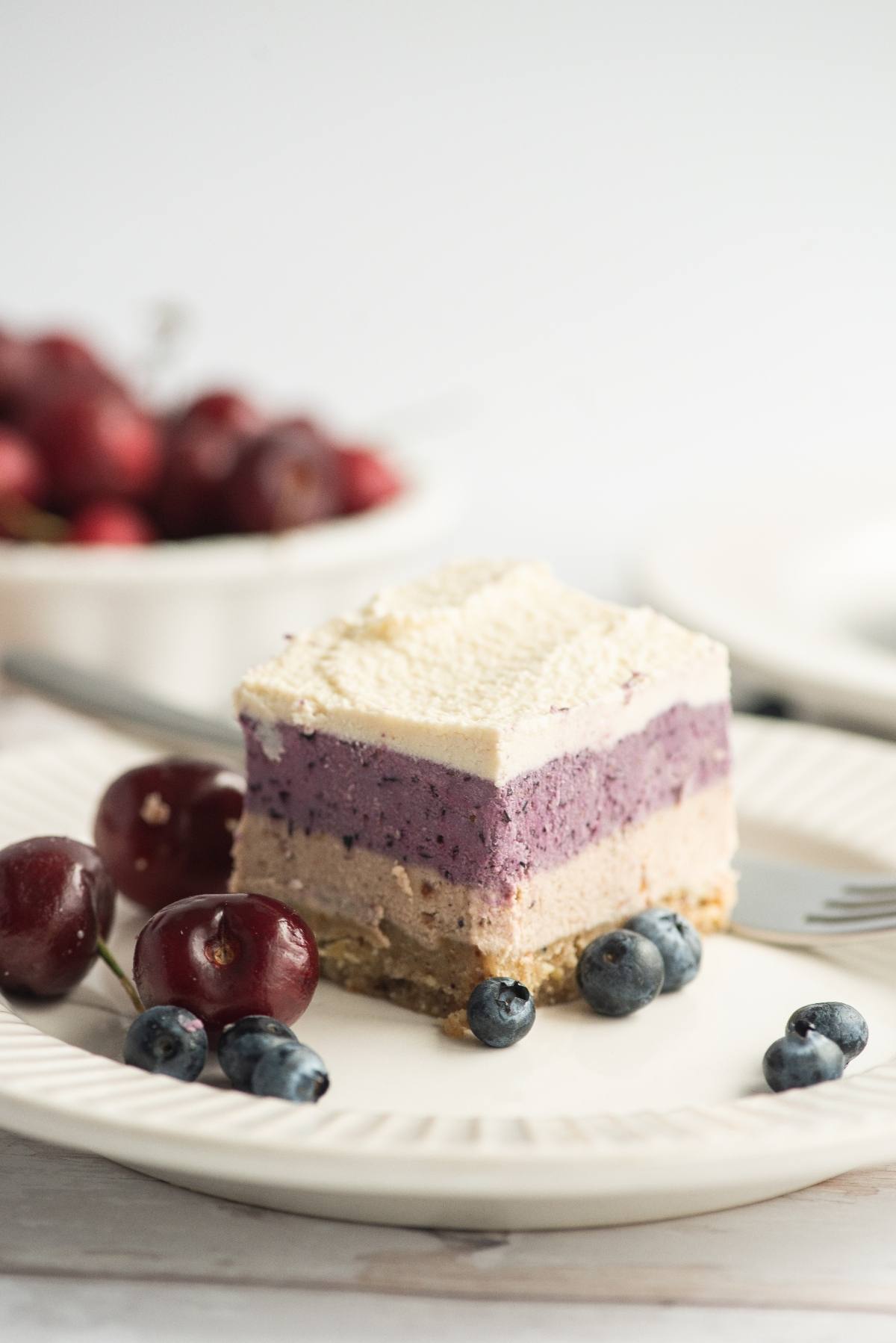 A slice of no bake vegan cheesecake, made with raw cashews and topped with blueberries and cherries, sits on a white plate.