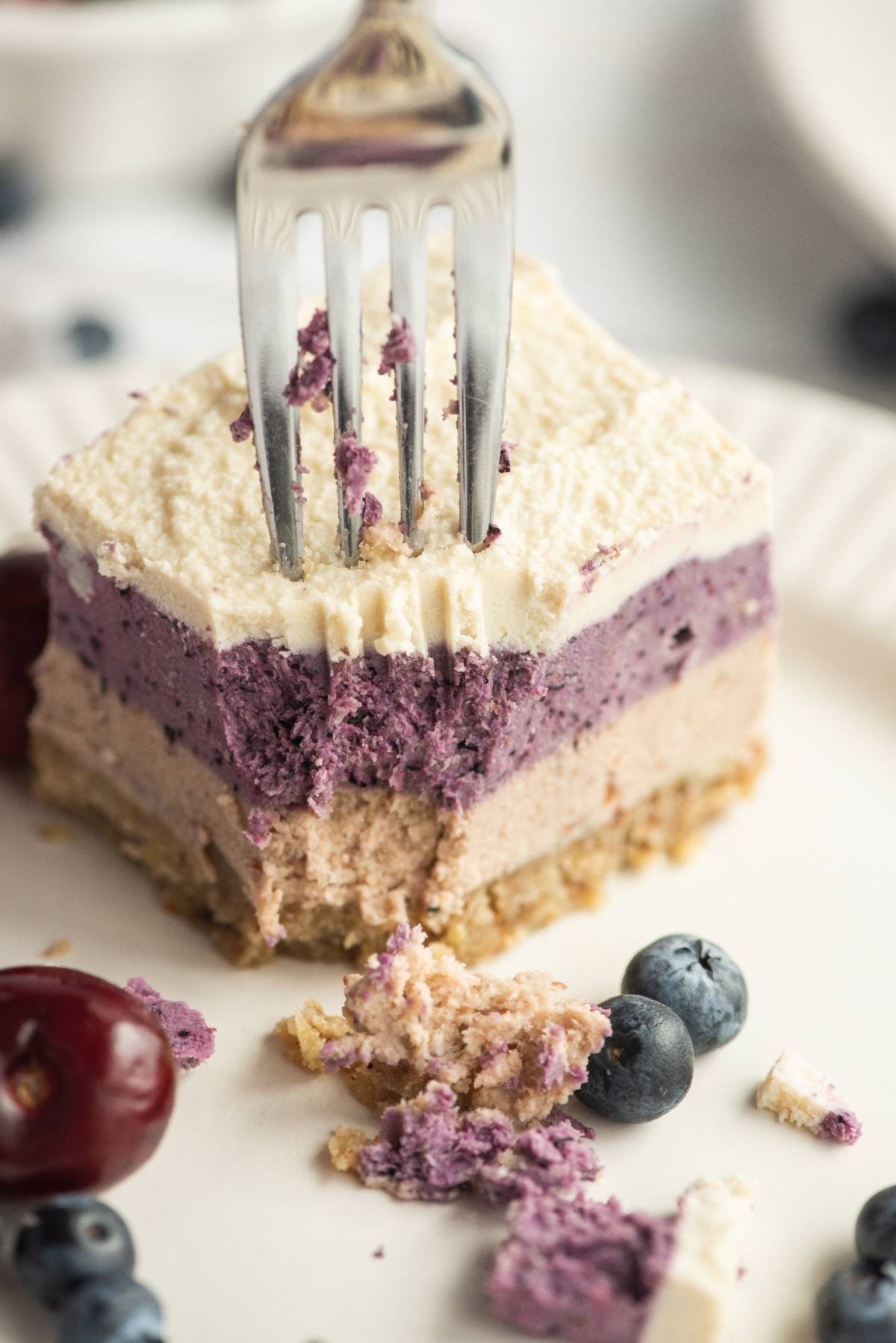 A fork slices into a layered cheesecake made with raw cashews and topped with blueberries and cherries on a white plate.