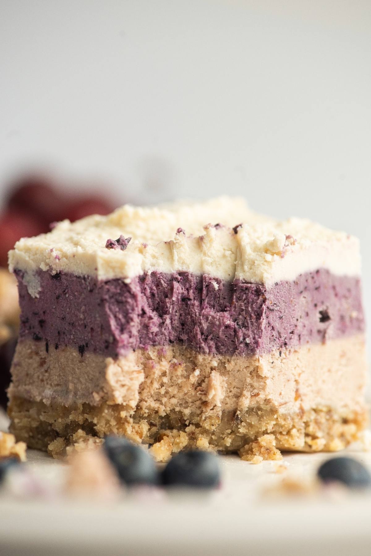 A no bake vegan cheesecake bar with a crumbly base and creamy purple and white layers, made with raw cashews and garnished with blueberries.