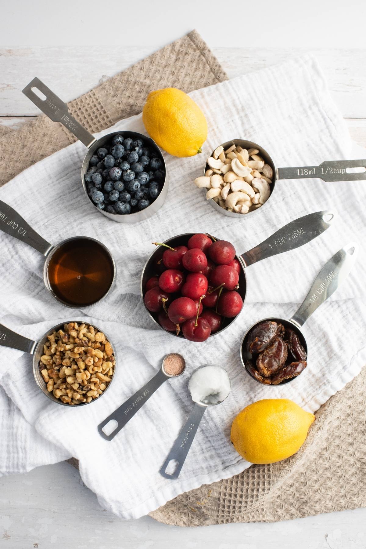 Overhead view of measuring cups with cherries, blueberries, nuts, dates, a lemon, and a small bowl of salt—perfect ingredients for a no bake vegan cheesecake made with raw cashews.