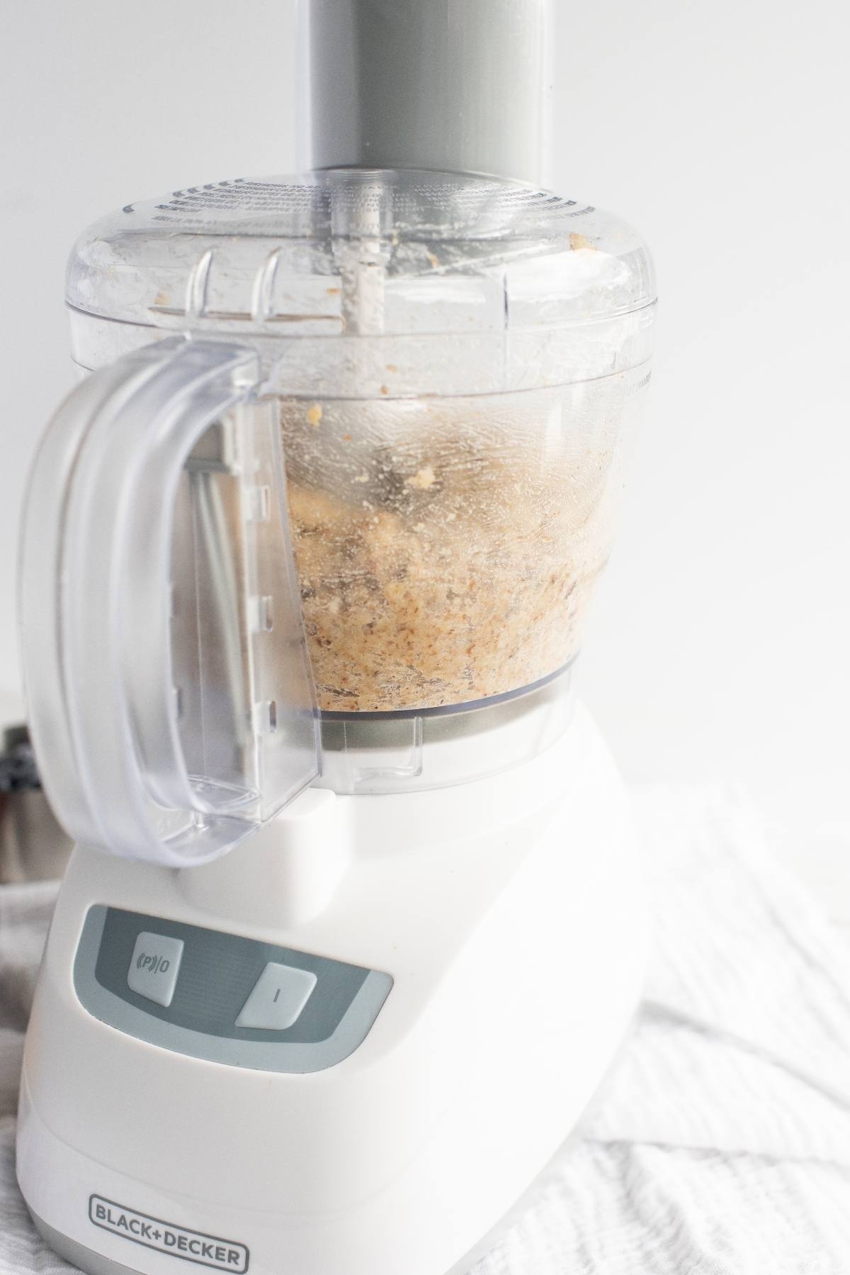 A food processor blending ingredients for a raw vegan recipe, with a creamy mixture made with raw cashews inside, on a white surface.