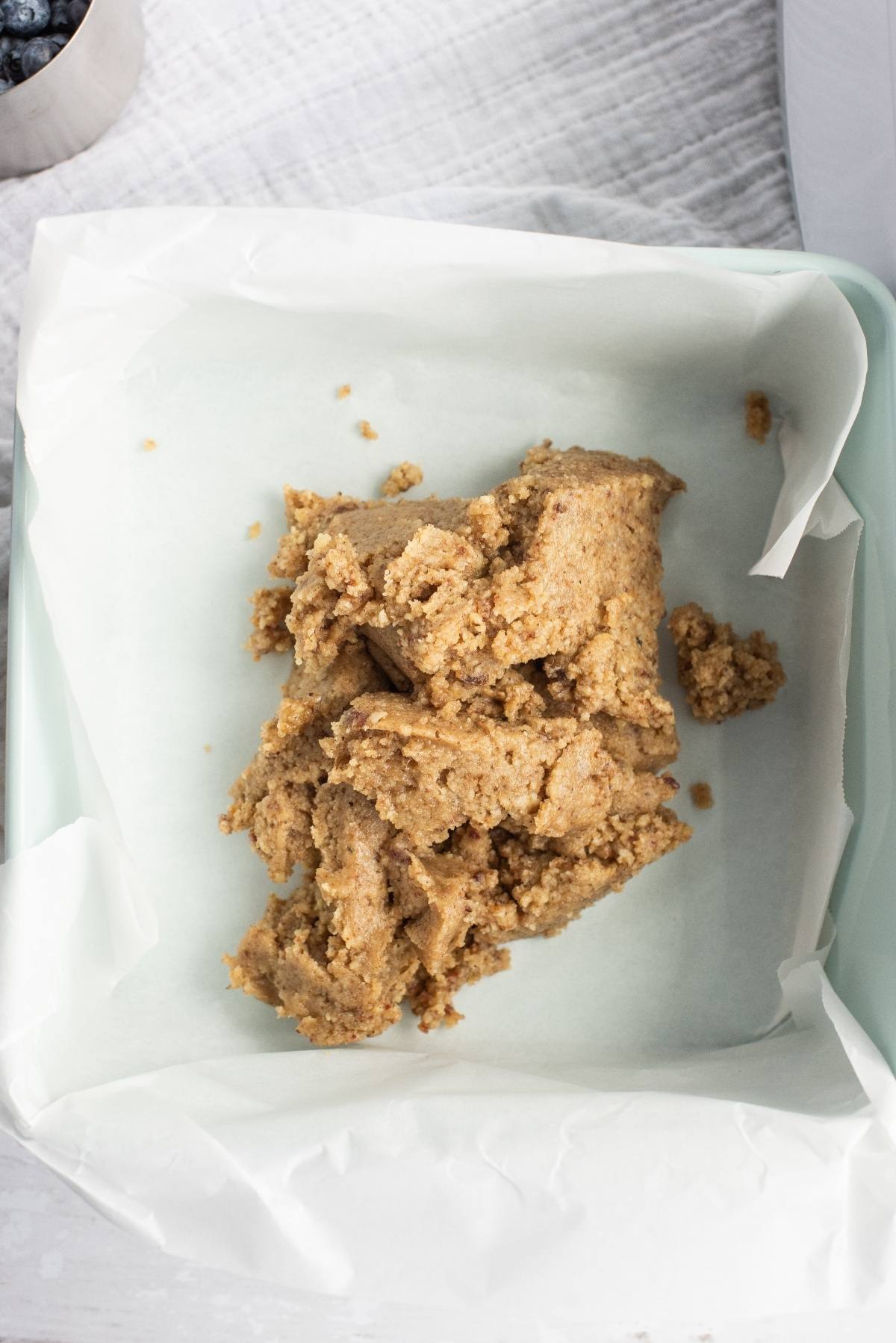 Brown dough mixture, made with raw cashews, placed in a parchment-lined square baking pan on a white surface—perfect for starting a vegan layered dessert.