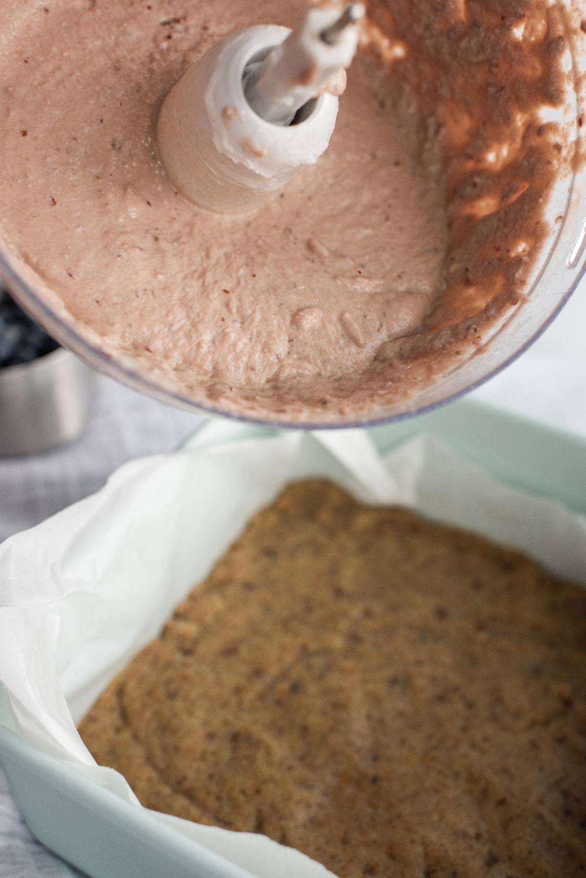 A blender with chocolate mixture held above a parchment-lined pan with a baked crust, perfect for creating a vegan layered dessert or a no bake vegan cheesecake made with raw cashews.