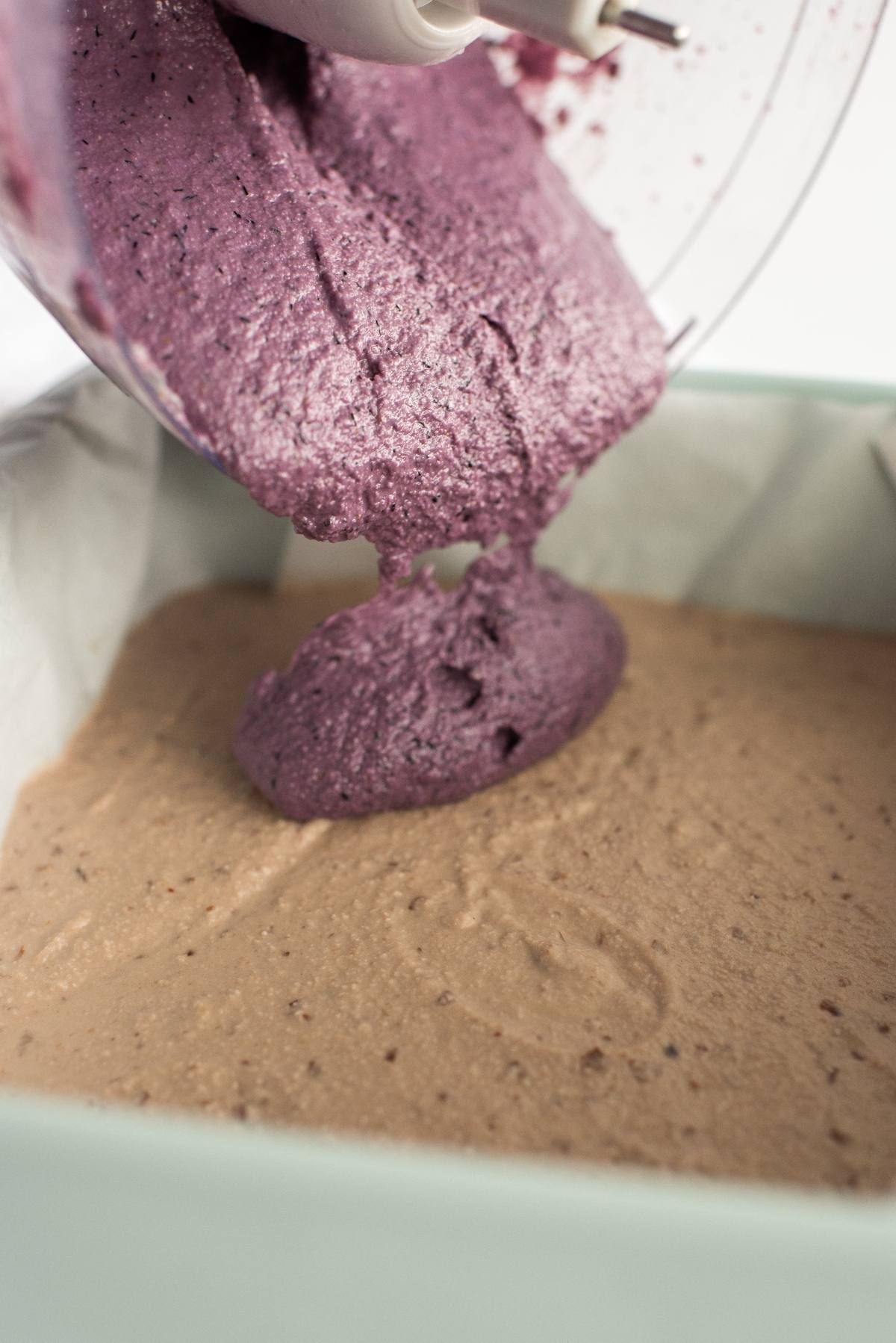 Thick purple batter for a no bake vegan cheesecake, made with raw cashews, is being poured from a mixer onto light brown batter in a parchment-lined pan.