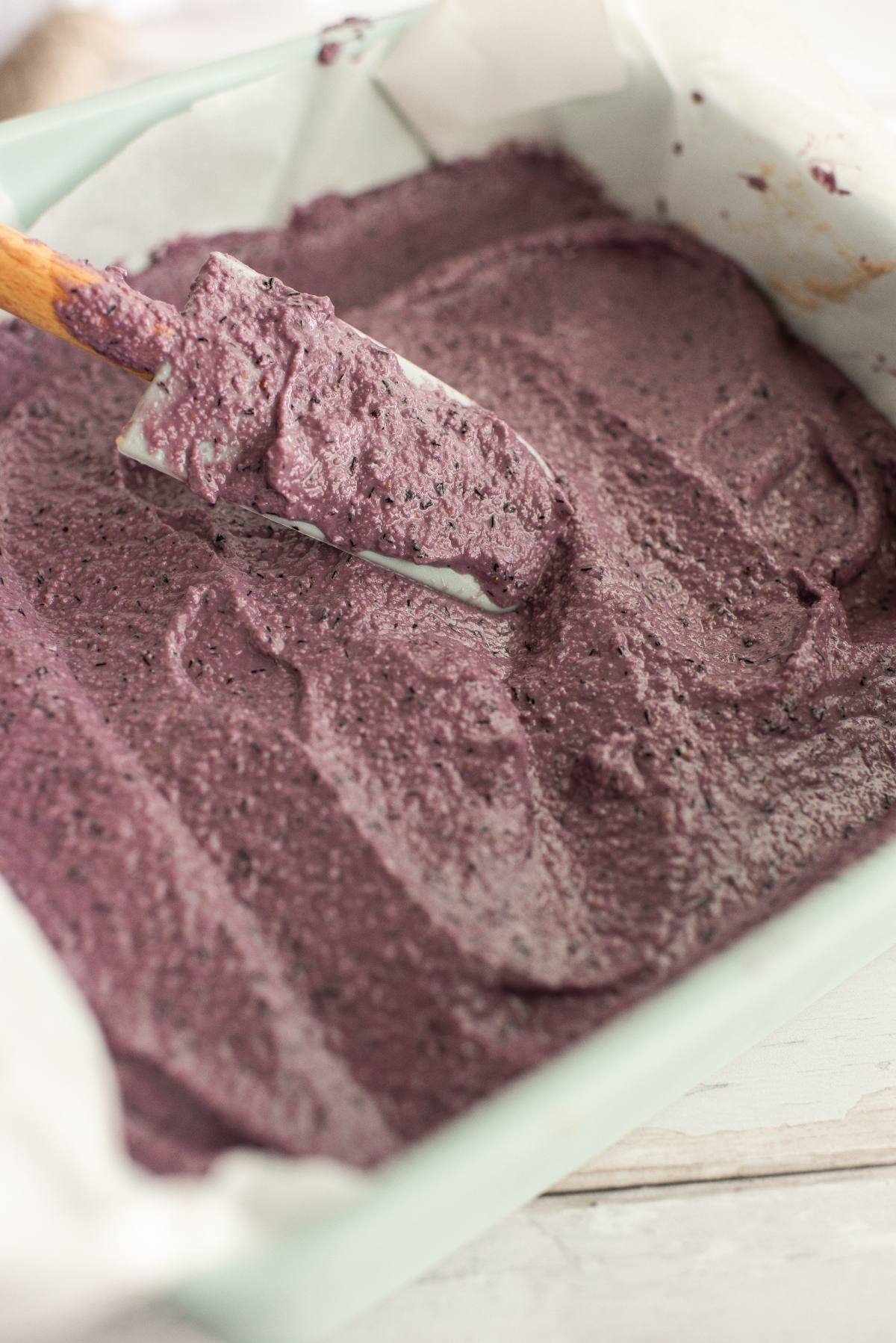 A spatula spreads thick, purple batter in a parchment-lined baking pan for a vegan layered dessert made with raw cashews.