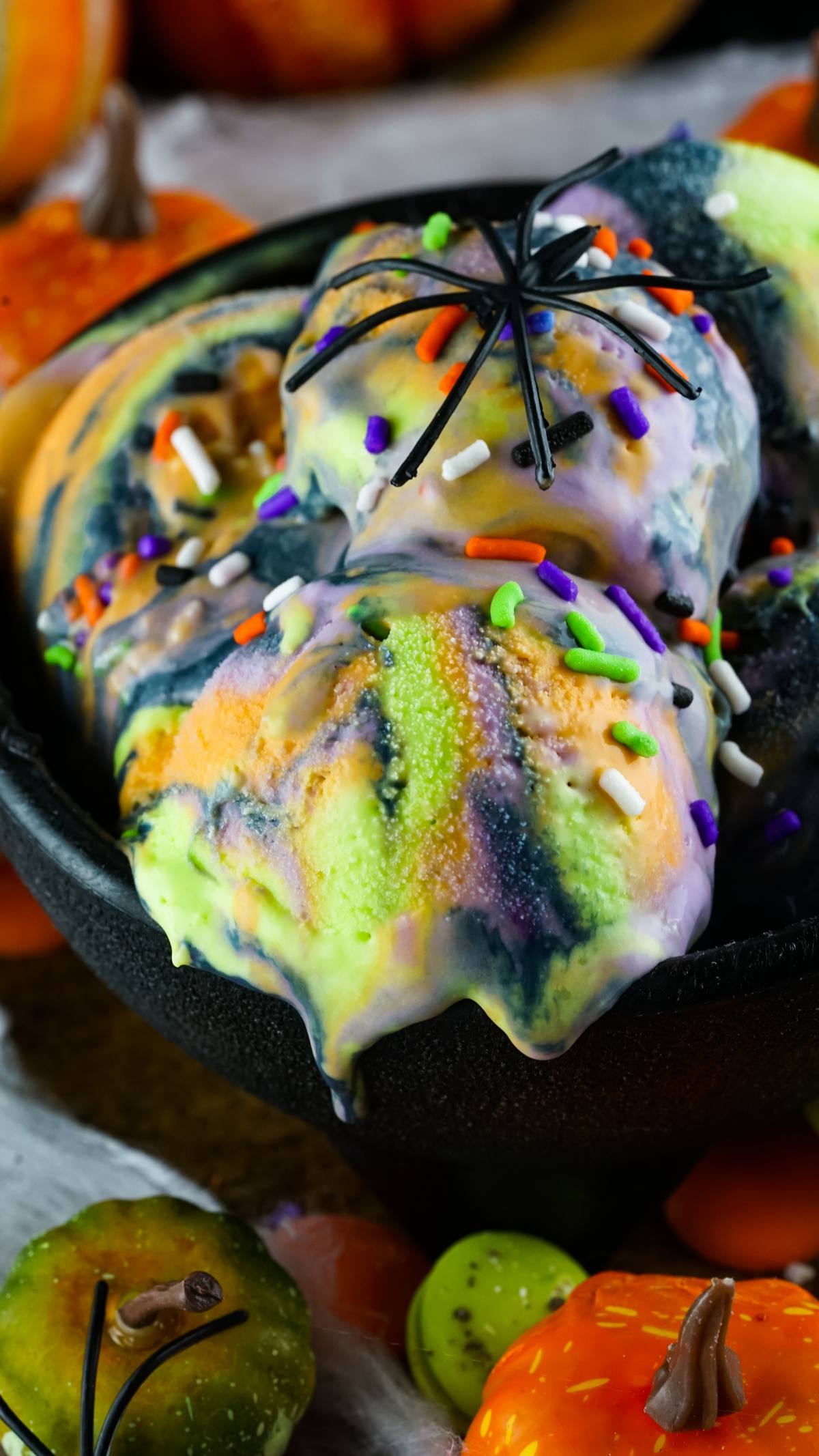 Colorful swirled spooky ice cream with Halloween sprinkles and a plastic spider in a black bowl, surrounded by mini pumpkins, makes the perfect frozen Halloween treat.