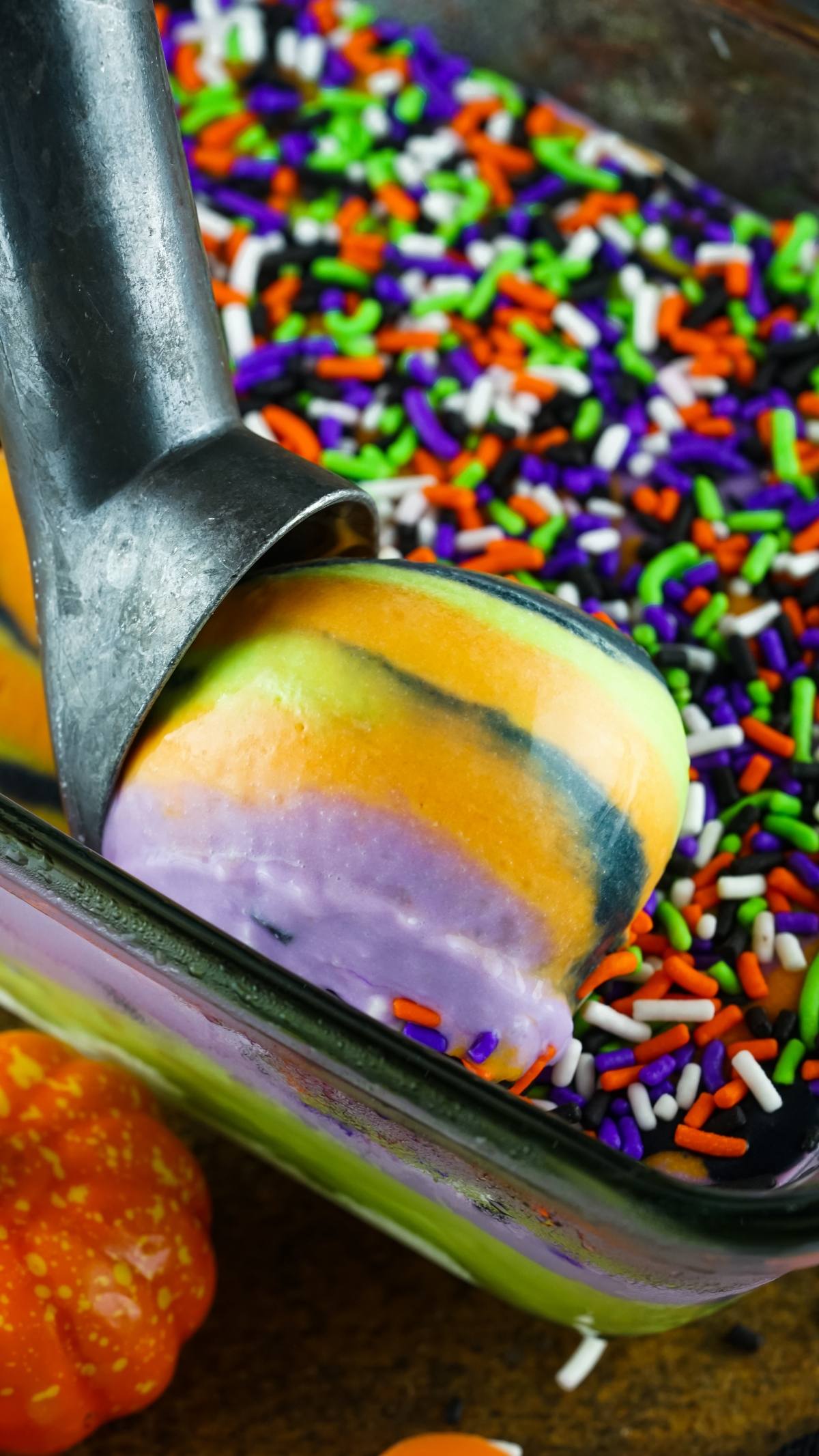 An ice cream scoop lifts rainbow-striped No Churn Halloween Ice Cream, covered in colorful sprinkles, from a glass dish—perfect for festive frozen Halloween treats.