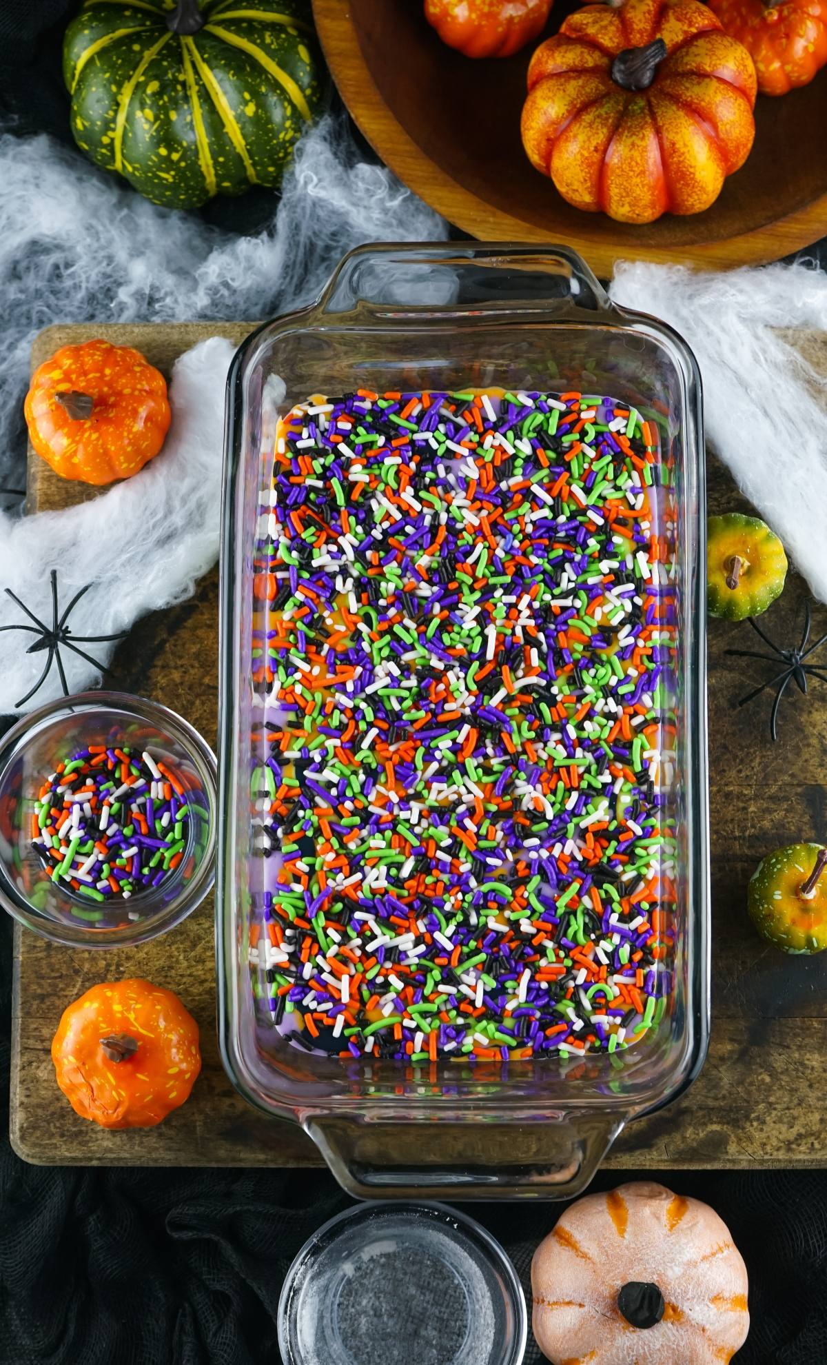 A glass dish filled with colorful Halloween sprinkles, surrounded by mini pumpkins and fake spiders, is the perfect setup for No Churn Halloween Ice Cream—one of the coolest frozen Halloween treats for your spooky celebration.