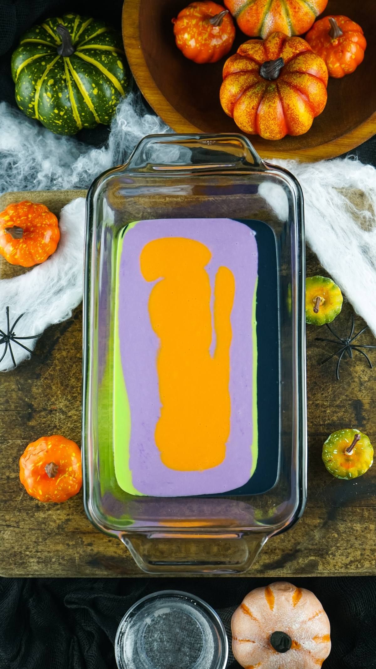 A glass dish with layered purple, orange, green, and black batter—this No Churn Halloween Ice Cream makes a festive dessert with Halloween colours, surrounded by pumpkins and spooky decor.