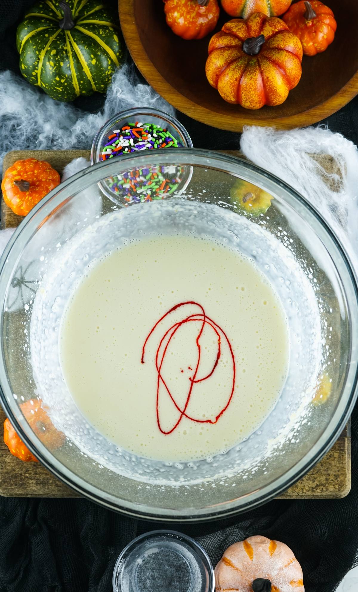 A glass bowl with batter and red food coloring, surrounded by pumpkins and Halloween sprinkles, sets the stage for making spooky ice cream or No Churn Halloween Ice Cream.