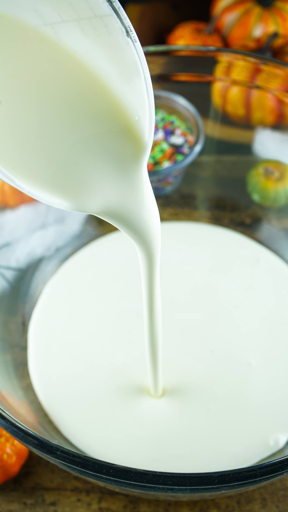 Cream being poured from a measuring cup into a glass bowl, surrounded by pumpkins and sprinkles—perfect for whipping up a No Churn Halloween Ice Cream or other frozen Halloween treats bursting with festive colours.