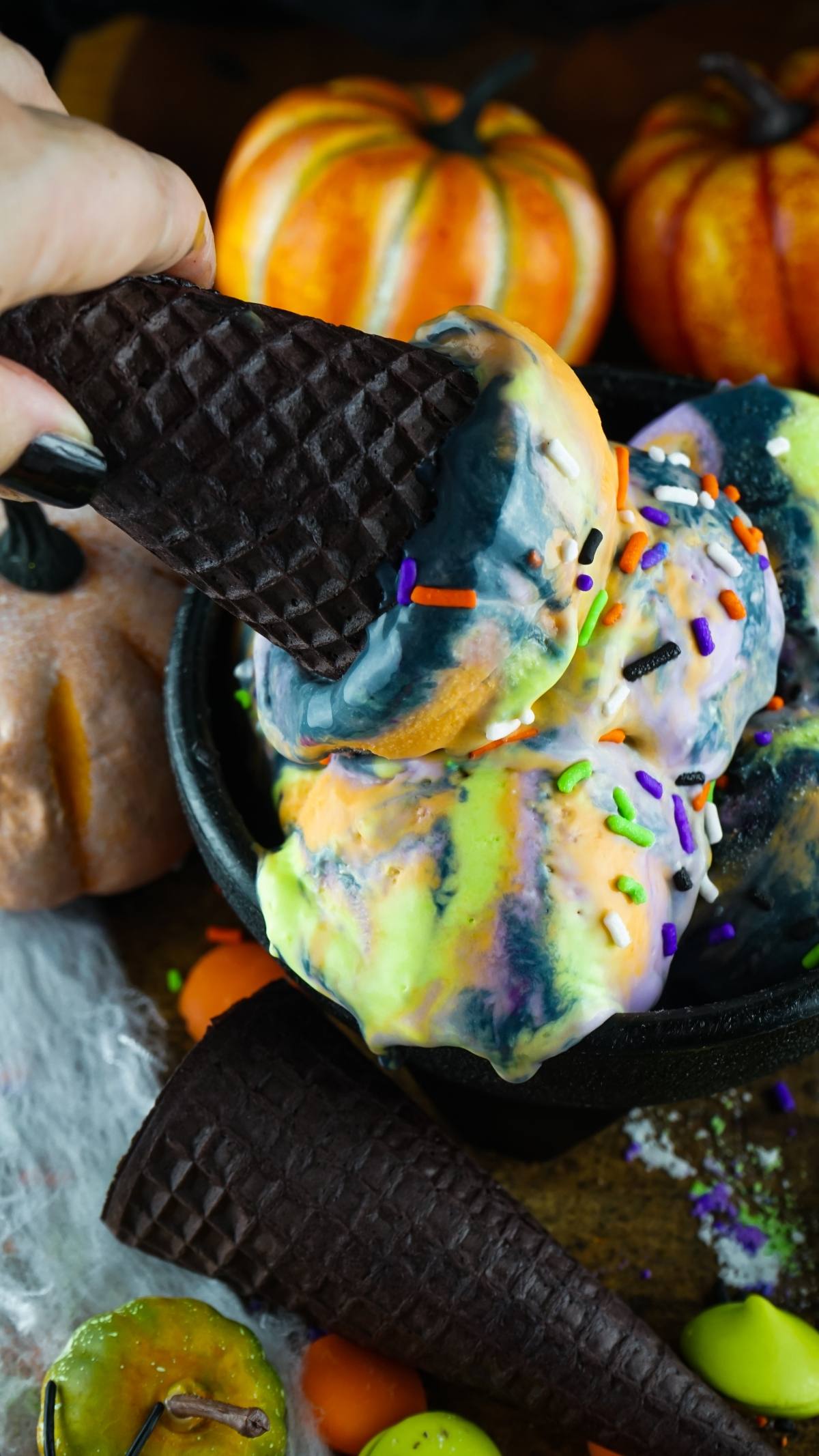 Hand holding a dark cone with birthday cake flavoured ice cream, topped with colorful sprinkles—a festive dessert with Halloween colours, surrounded by pumpkins and spooky decor.