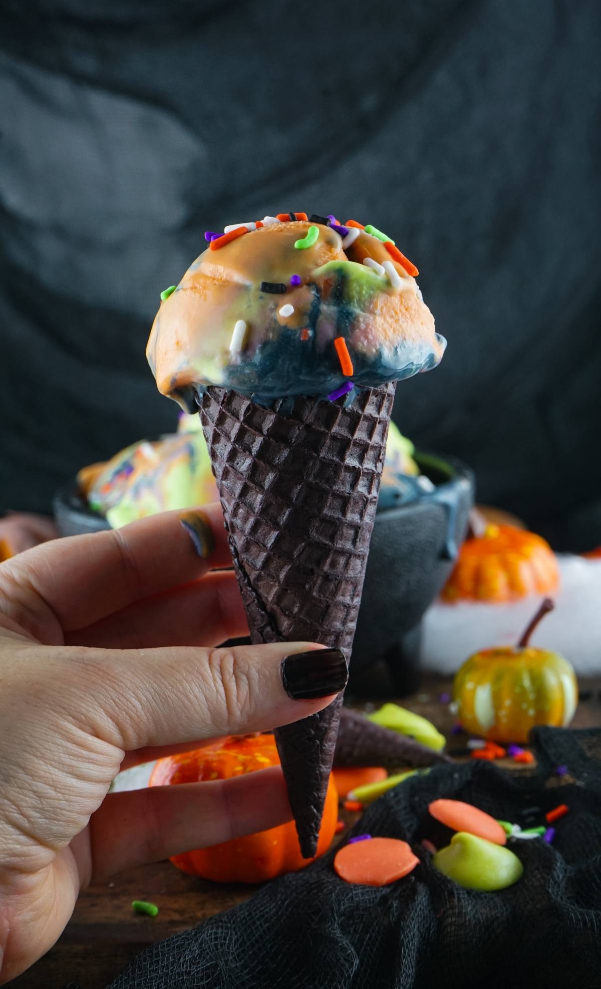 Hand holding a dark cone with orange, green, and black spooky ice cream topped with colorful sprinkles—a delicious dessert with Halloween colours.