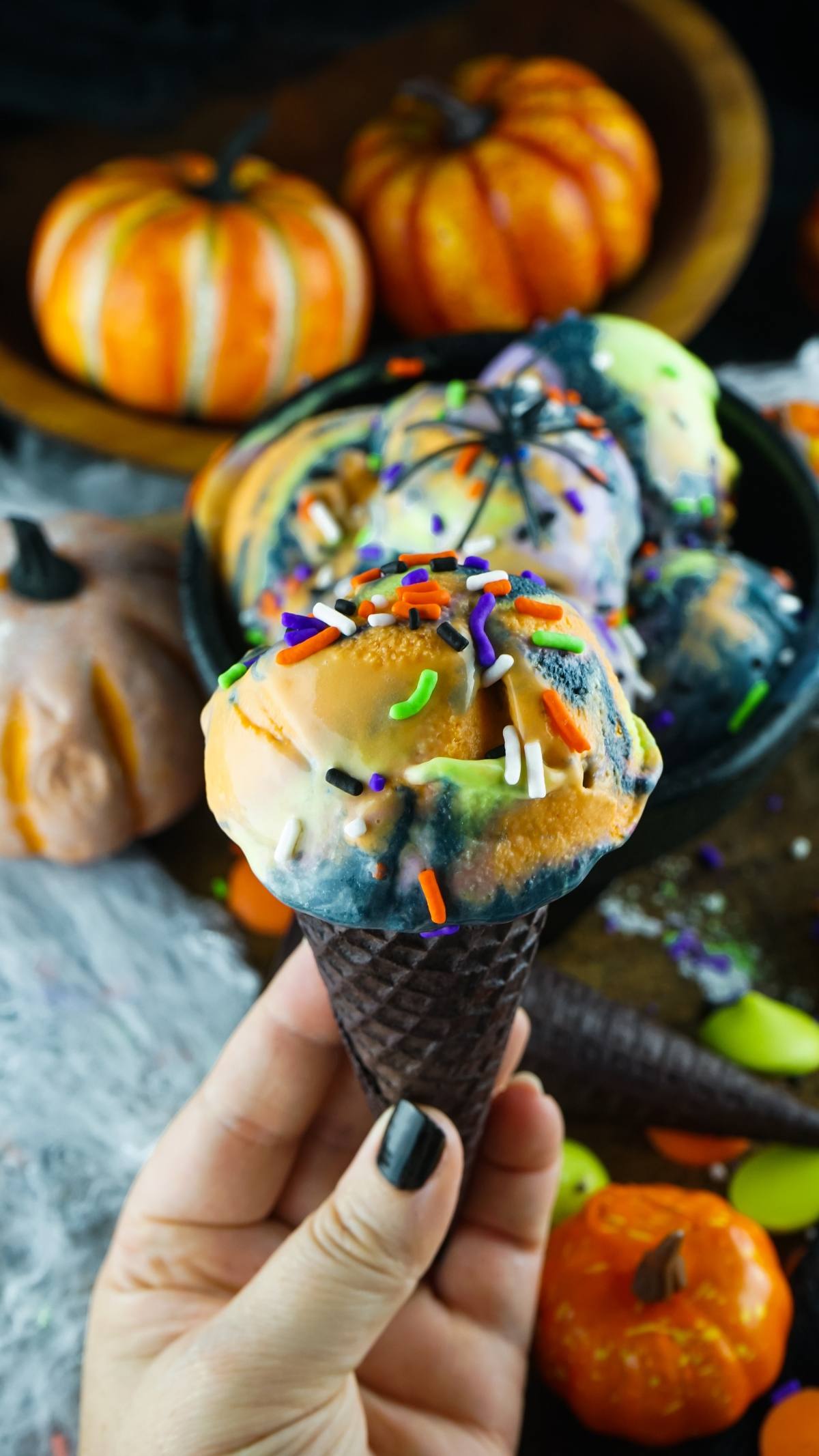 Hand holding a black cone with birthday cake flavoured ice cream in orange, purple, and black—spooky ice cream topped with Halloween sprinkles.