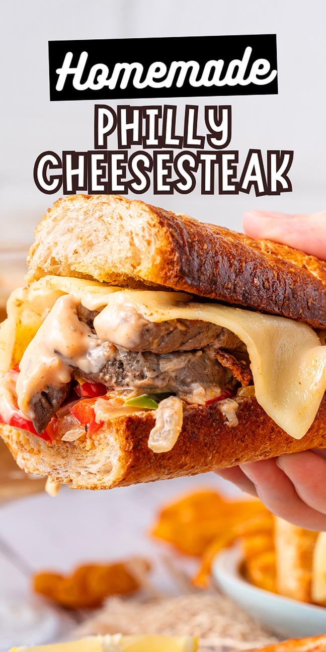 A hand holding a homemade Philly Cheesesteak sandwich loaded with melted cheese and savory beef on a crusty roll.