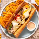 A Philly Cheesesteak sandwich with melted cheese, waffle fries, and dipping sauce served on a plate.