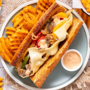 A Philly Cheesesteak sandwich with melted cheese, waffle fries, and dipping sauce served on a plate.