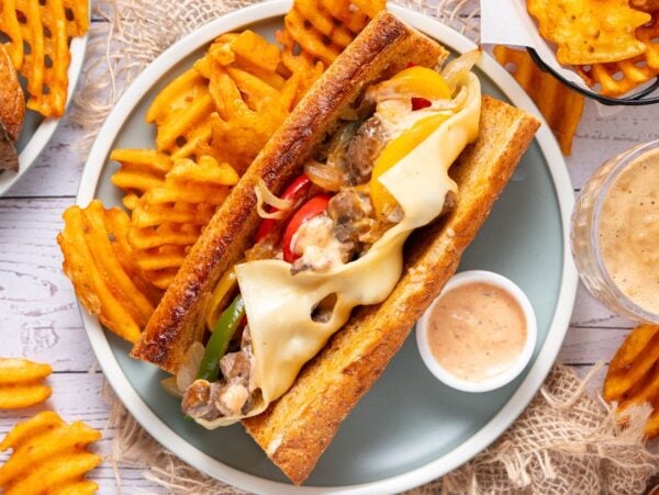 A Philly Cheesesteak sandwich with melted cheese, waffle fries, and dipping sauce served on a plate.
