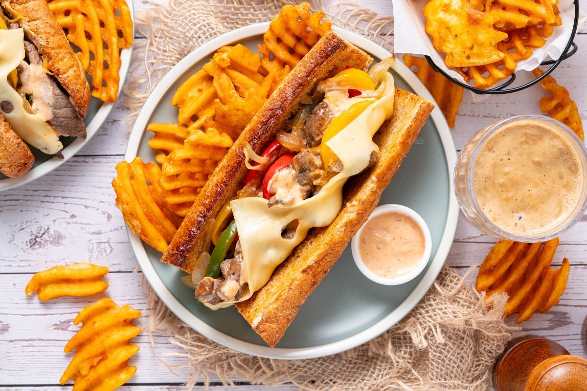 Enjoy a classic Philly Cheesesteak with melted cheese, served alongside crispy waffle fries and savory dipping sauce—all perfectly paired with a refreshing drink nearby.