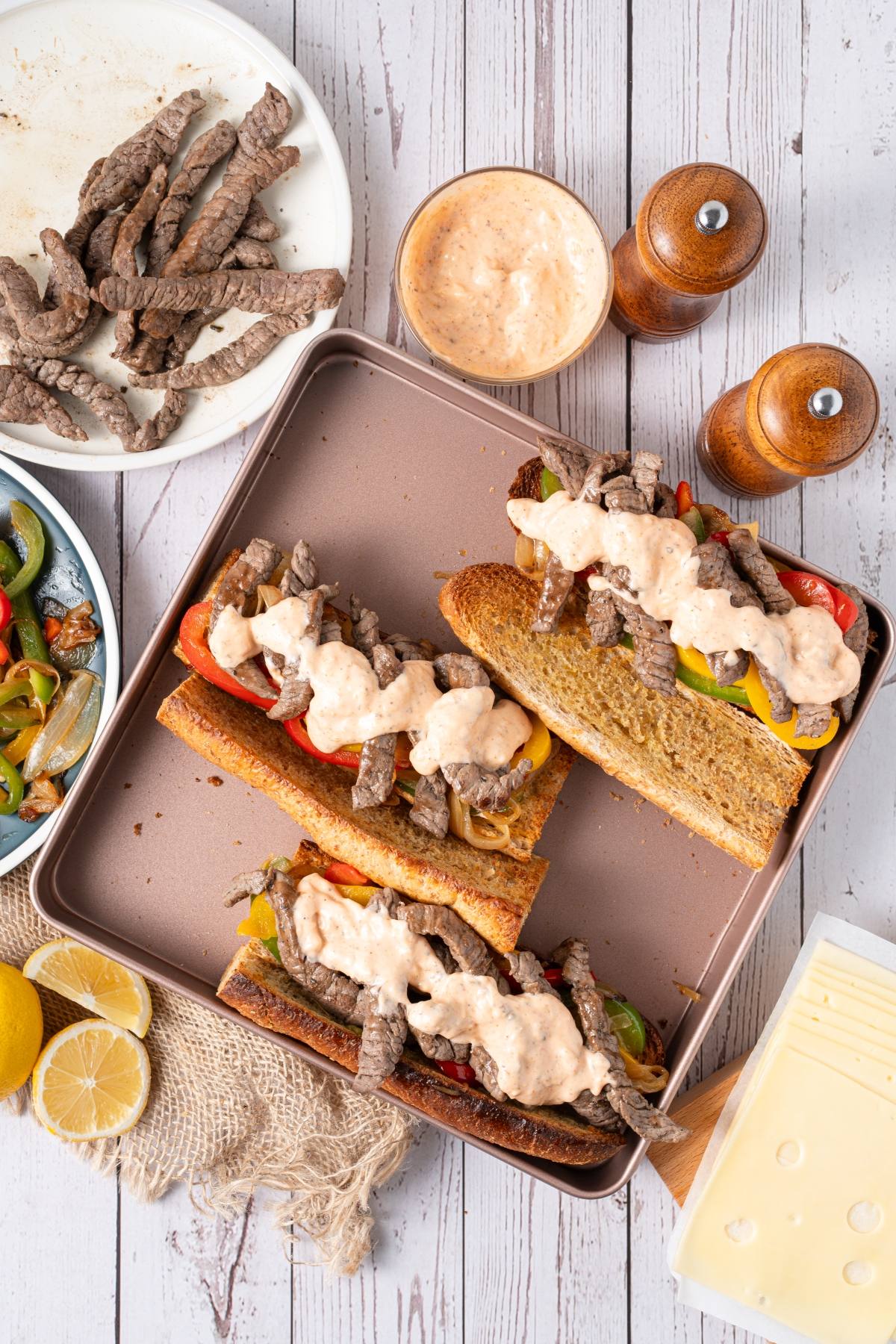 Three steak subs topped with sauce, surrounded by sliced steak, veggies, cheese, and condiments on a table.