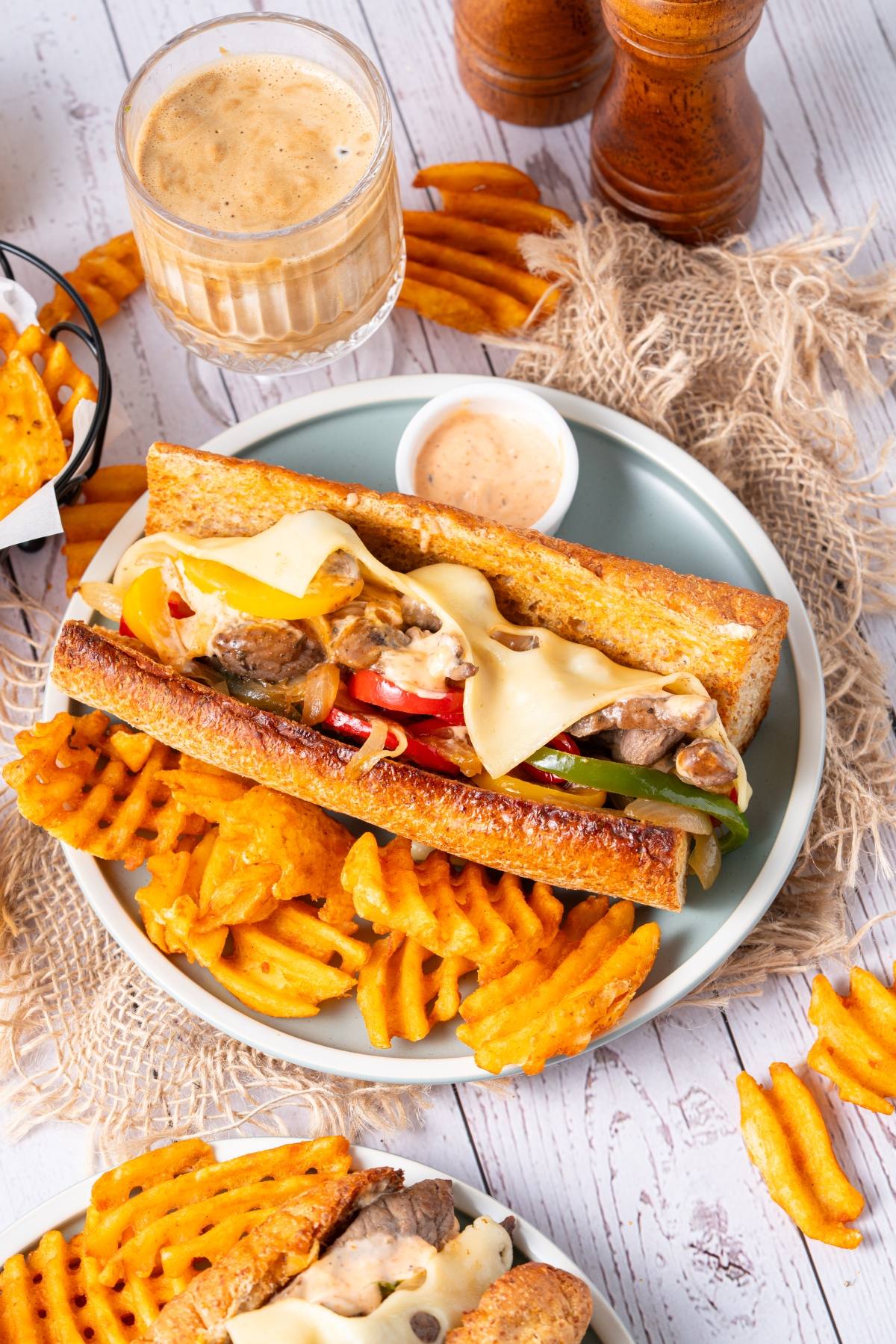 A sandwich with melted cheese, steak, and peppers, served with waffle fries and a dipping sauce.