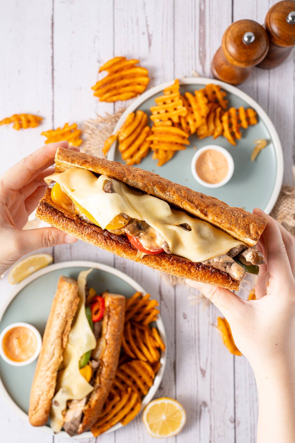 A person holds a cheesy sub sandwich above a plate of waffle fries and dipping sauce.