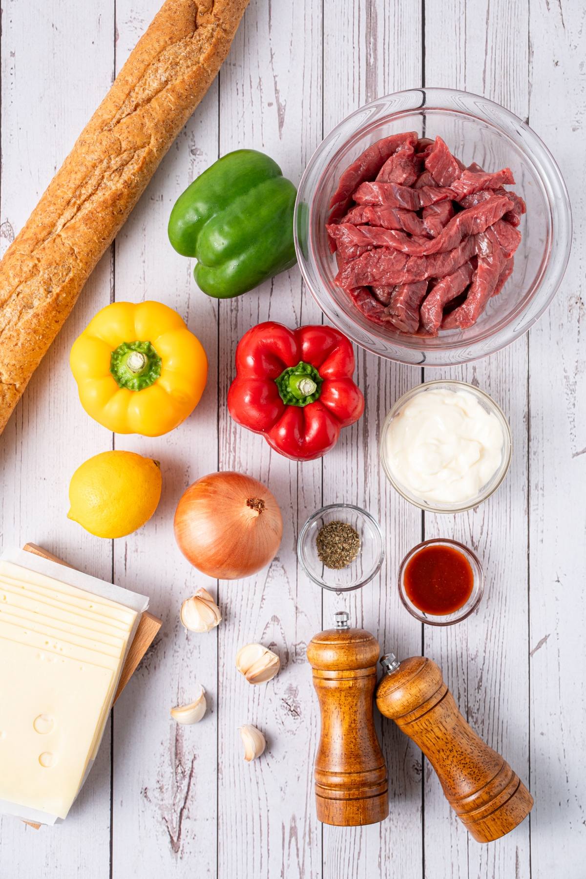 Sliced beef, bell peppers, onion, cheese, baguette, garlic, spices, lemon, and sauces on a white wood table.