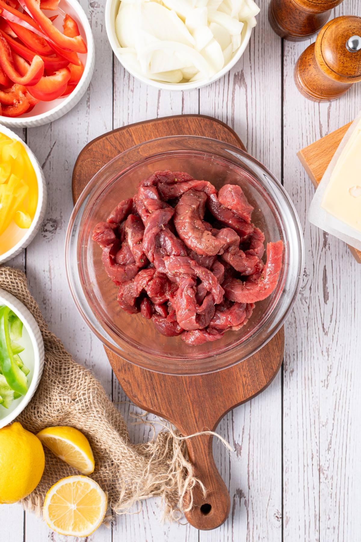 Raw beef strips in a glass bowl surrounded by sliced peppers, onions, lemon halves, and seasoning on a wooden table.