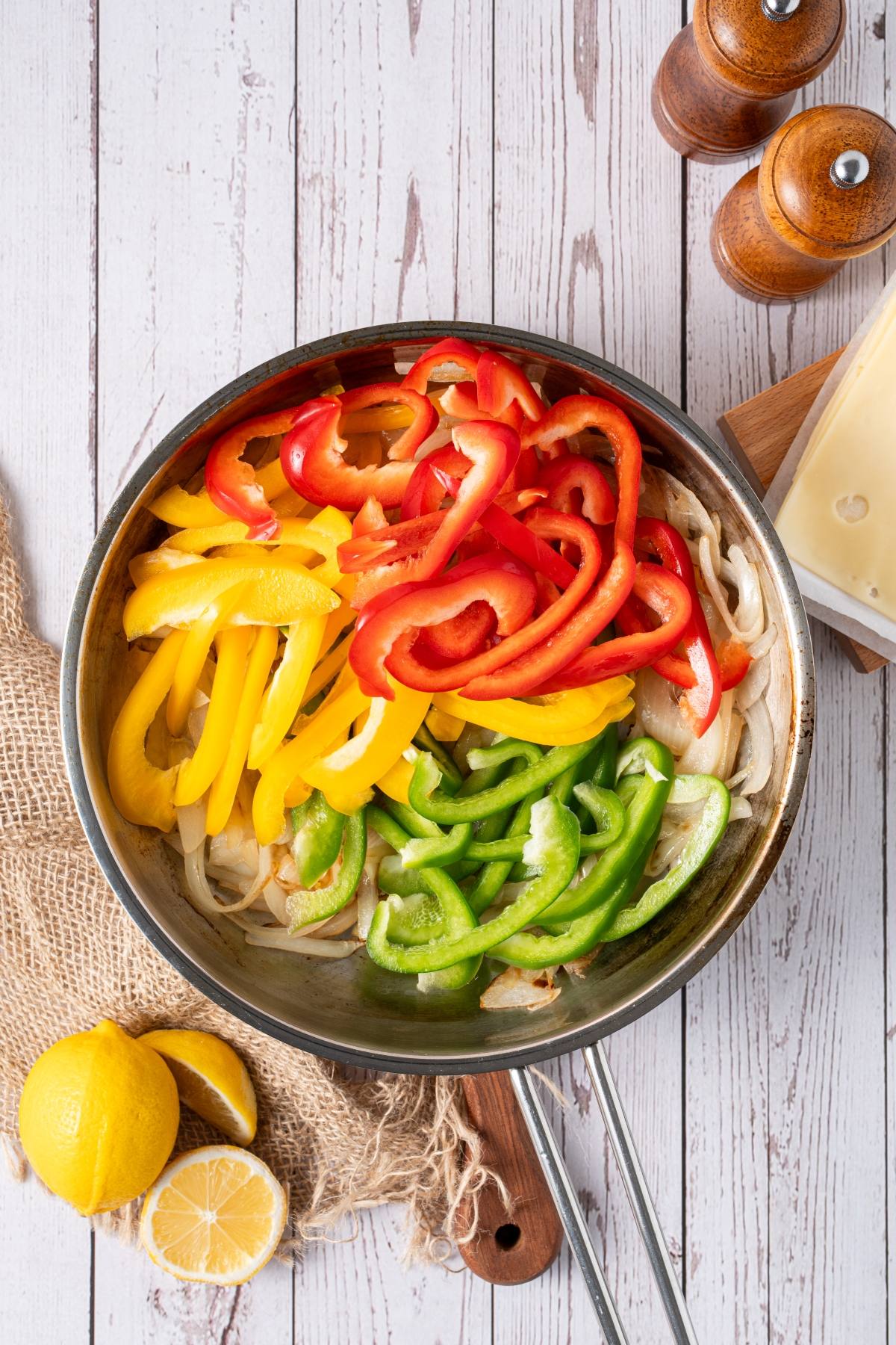 A skillet with sliced red, yellow, and green bell peppers and onions on a white wooden table.