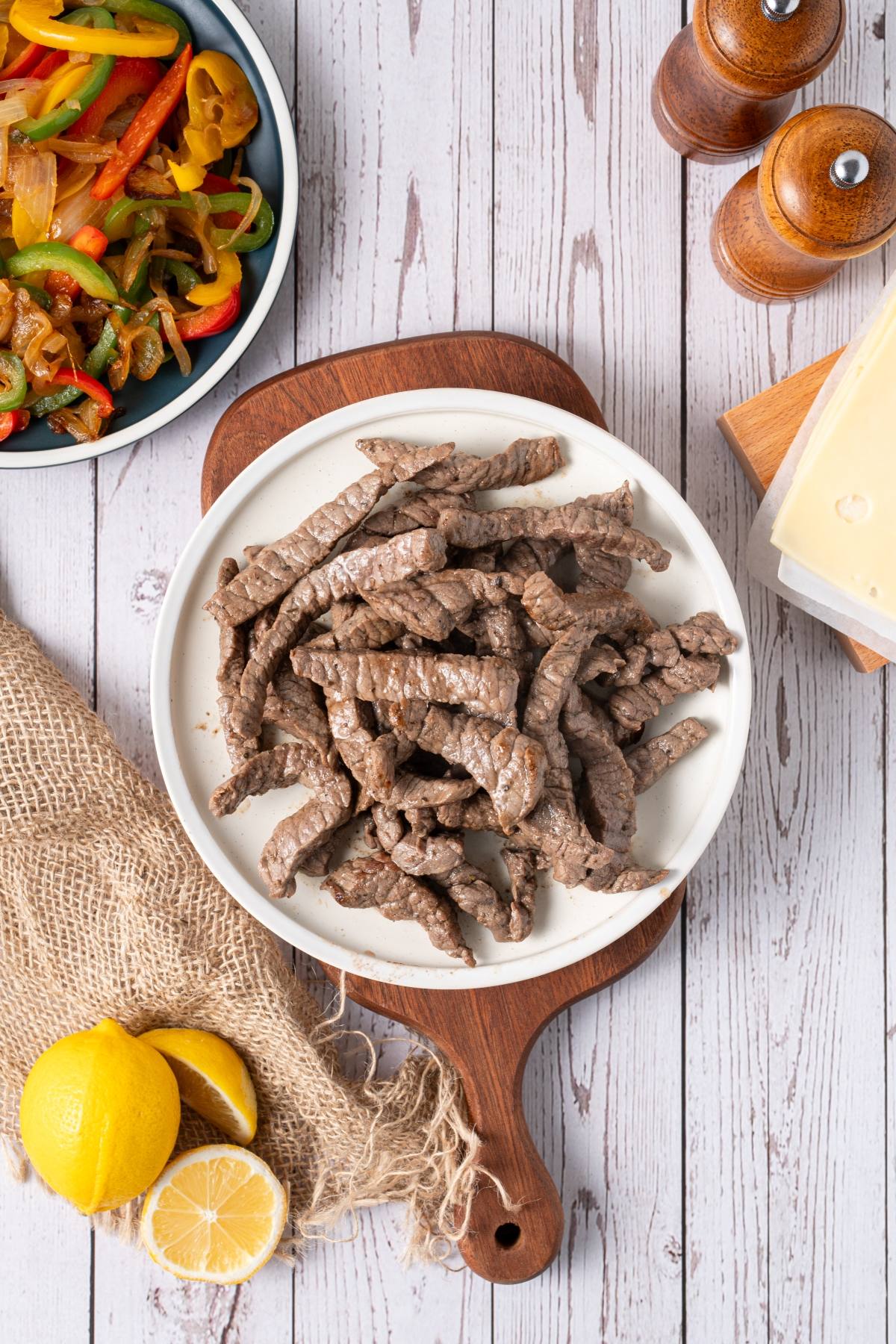 Sliced cooked beef on a white plate, with sautéed peppers, lemons, cheese, and pepper mills nearby.