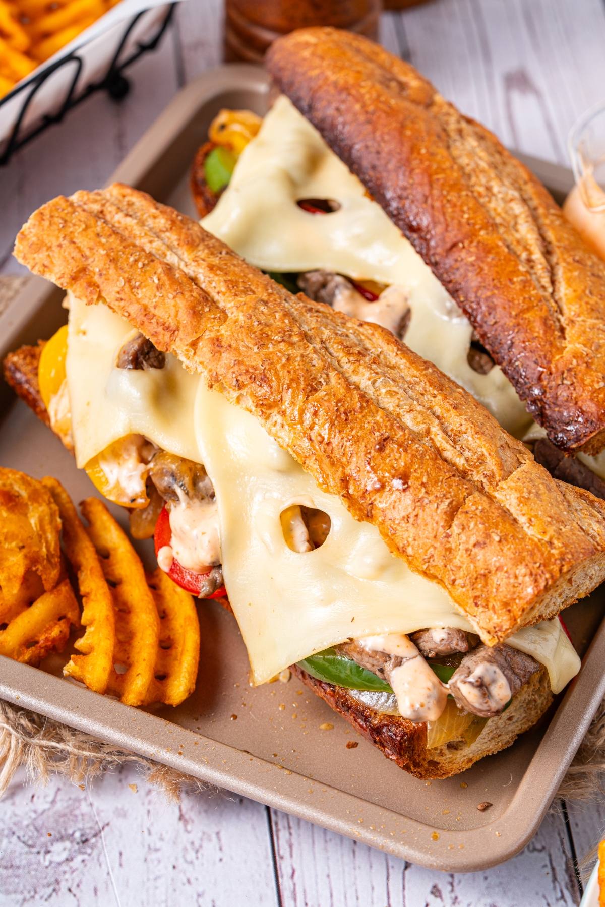 Two sub sandwiches with melted cheese, mushrooms, and veggies on a tray with waffle fries on the side.