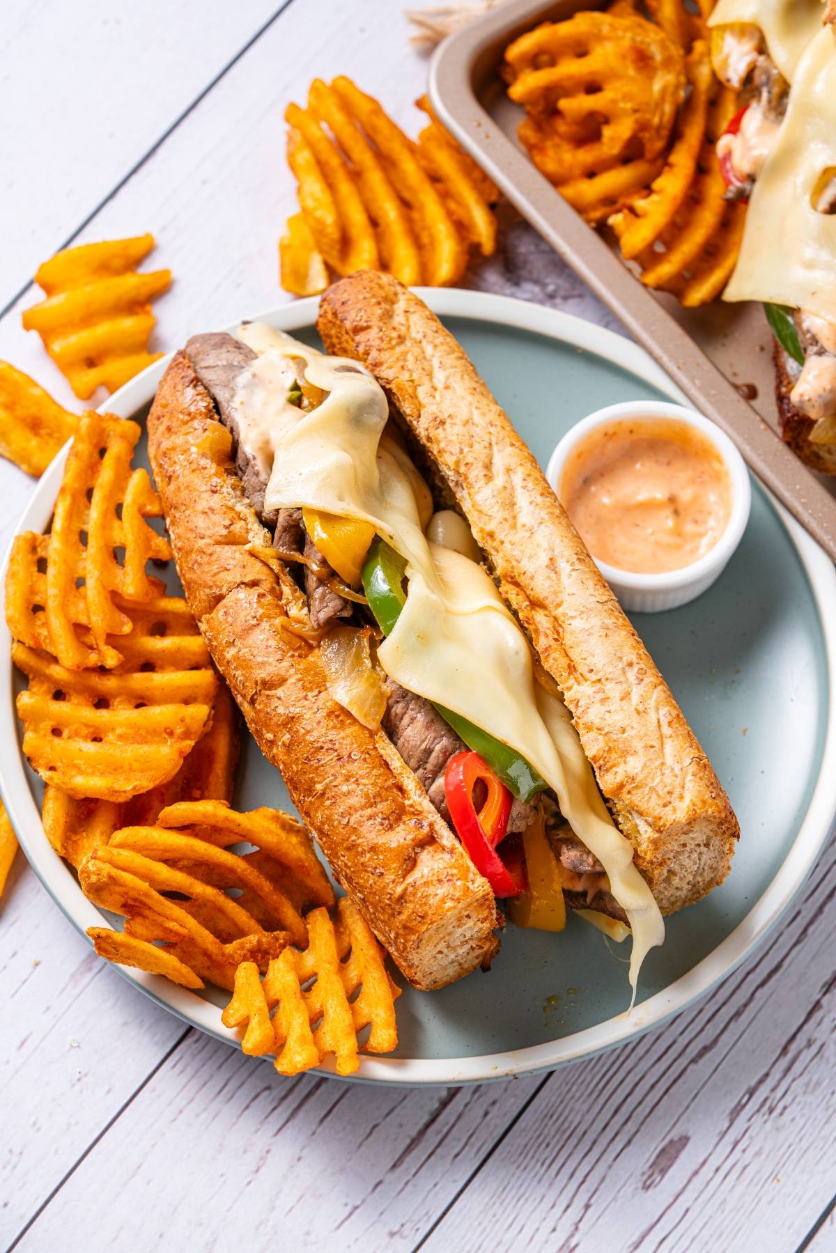 Philly cheesesteak sandwich with melted cheese and waffle fries on a plate, with dipping sauce on the side.