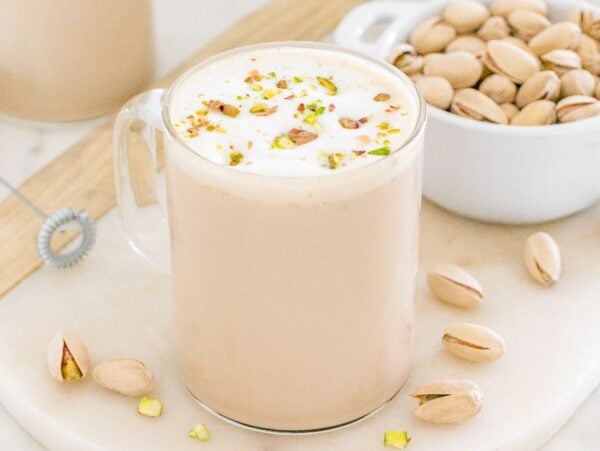 A mug of creamy pistachio latte topped with foam and chopped pistachios sits beside a bowl of whole pistachios.