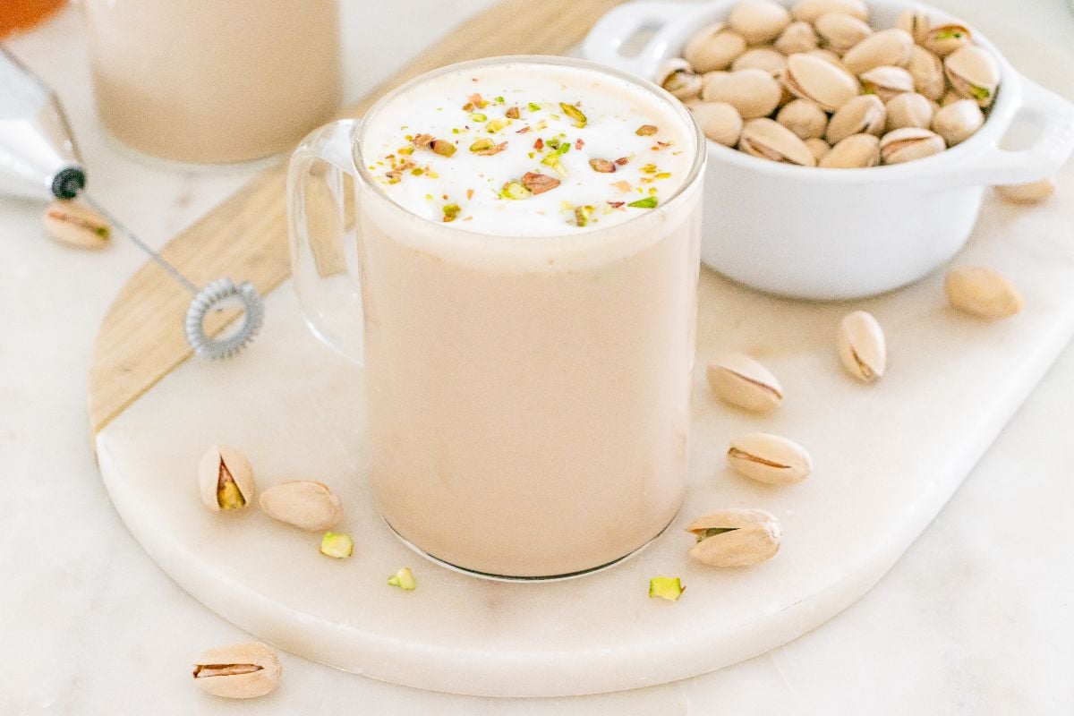 A glass mug of pistachio latte topped with creamy foam and crushed pistachios, served beside a bowl of whole pistachios.