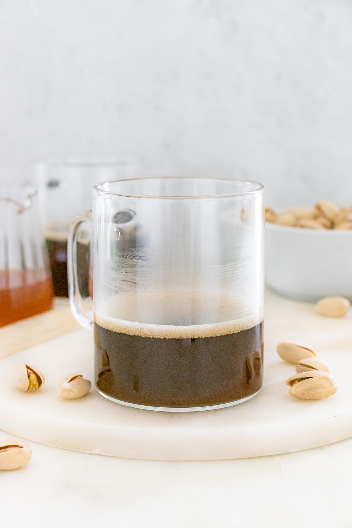 A clear glass mug of black coffee sits on a table with pistachios and a bowl in the background.
