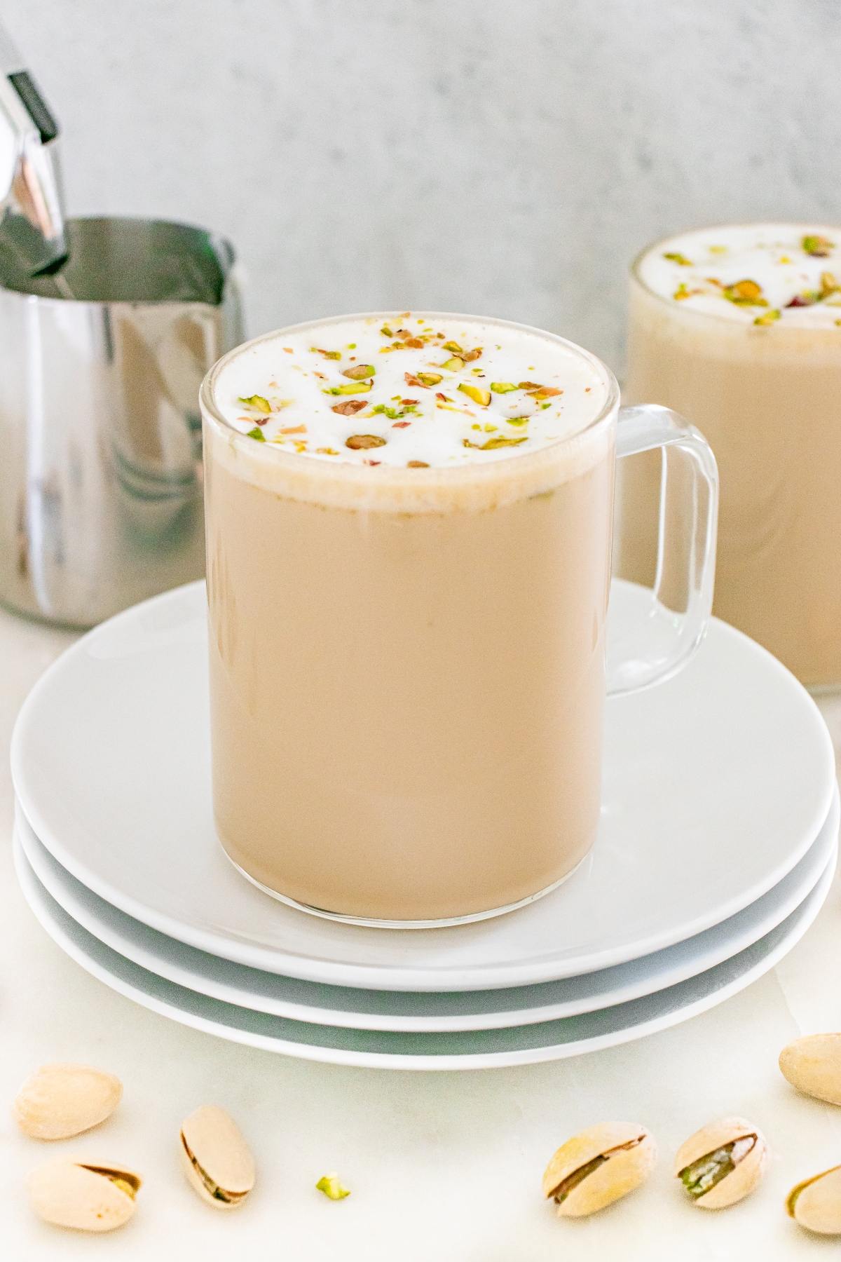 A glass mug of chai topped with foam and chopped pistachios, on stacked white plates with pistachios nearby.