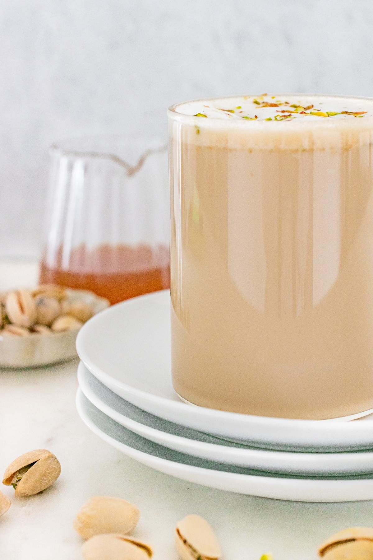 A glass of pistachio latte on stacked white plates, with pistachios and syrup in the background.