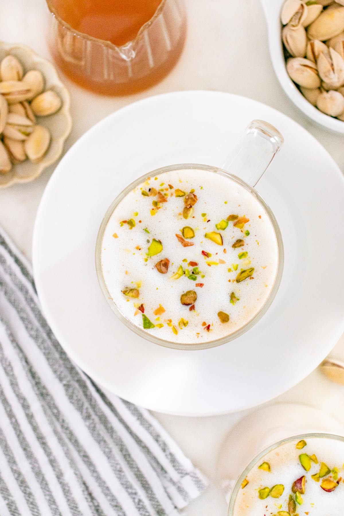 A glass mug of frothy milk topped with chopped pistachios, next to bowls of pistachios and a striped towel.