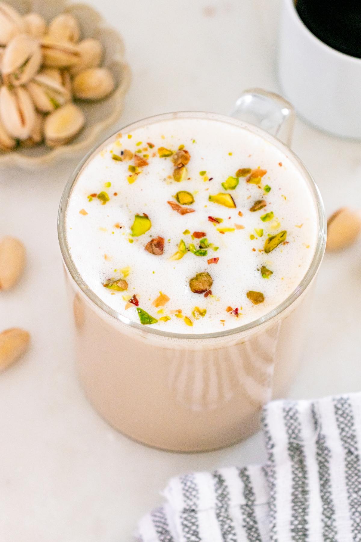 Glass mug of foamy latte topped with crushed pistachios, next to pistachios and a striped napkin.